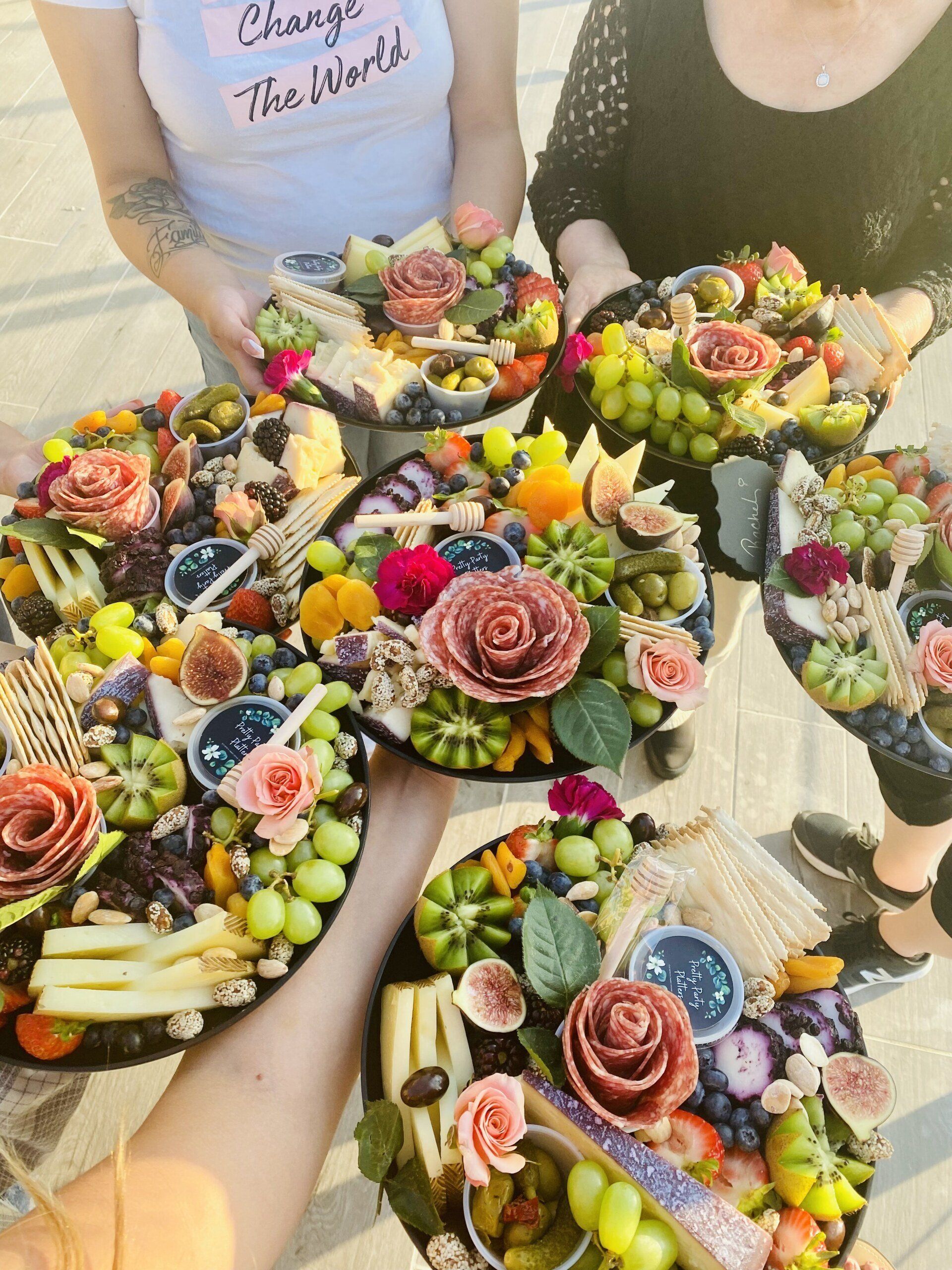 Five people hold colorful charcuterie boards outdoors. Boards feature fruit, cheese, and decorative flowers.