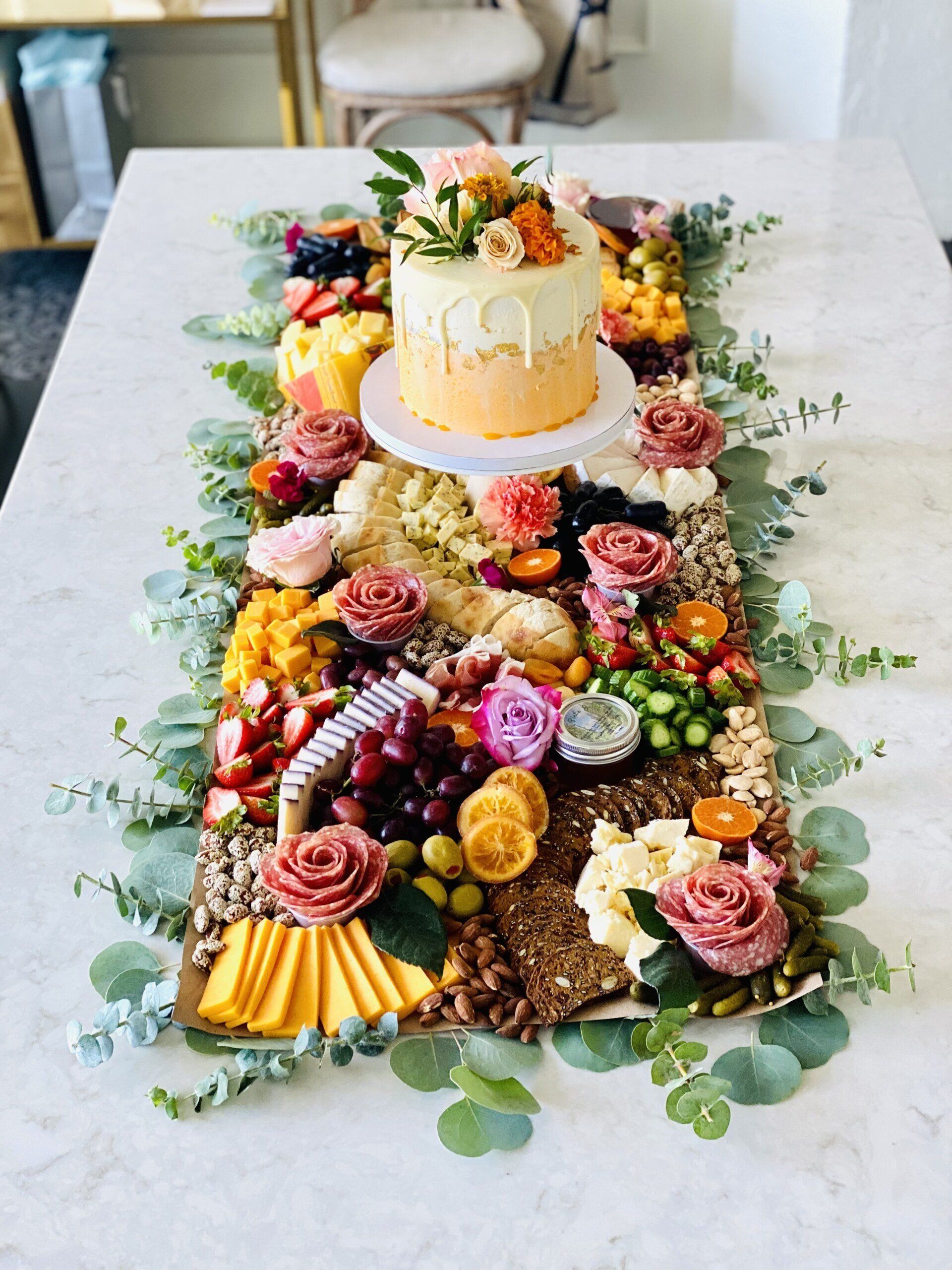 Charcuterie board with cake, colorful fruits, flowers, cheeses, and greenery on a white table.