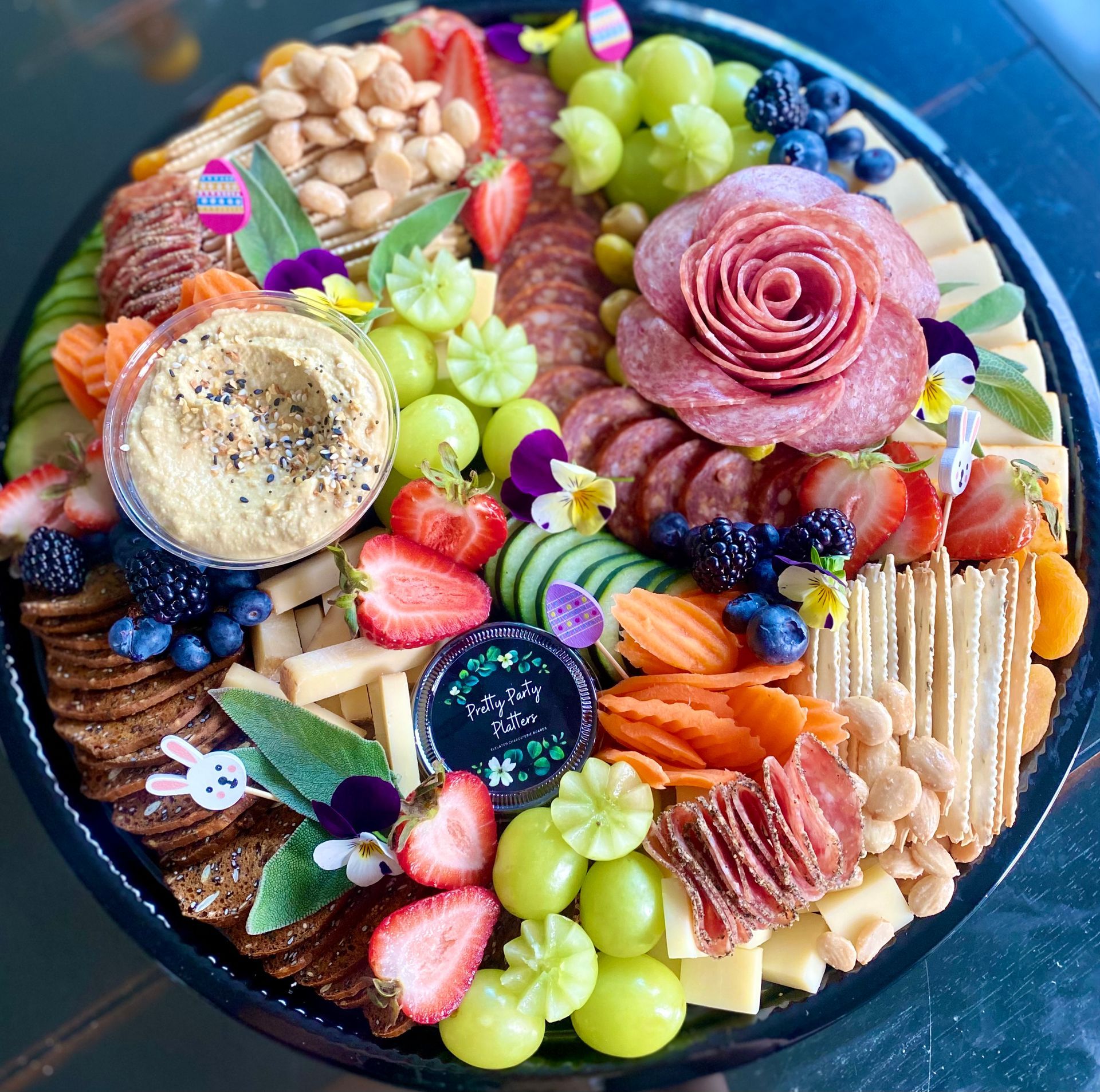 Charcuterie board with salami rose, hummus, fruits, crackers, nuts, cheeses, and garnishes.