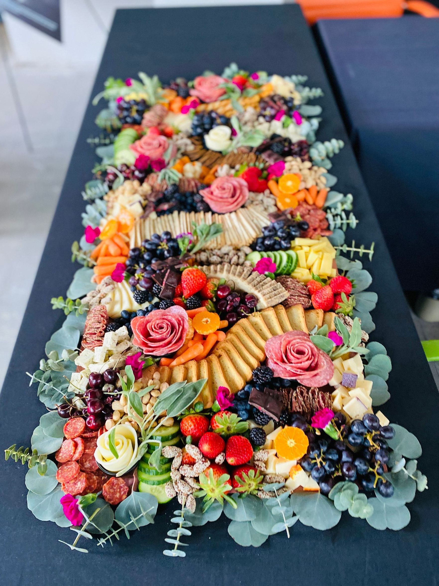 Long charcuterie board with cheese, crackers, fruit, flowers, and greenery on a black surface.