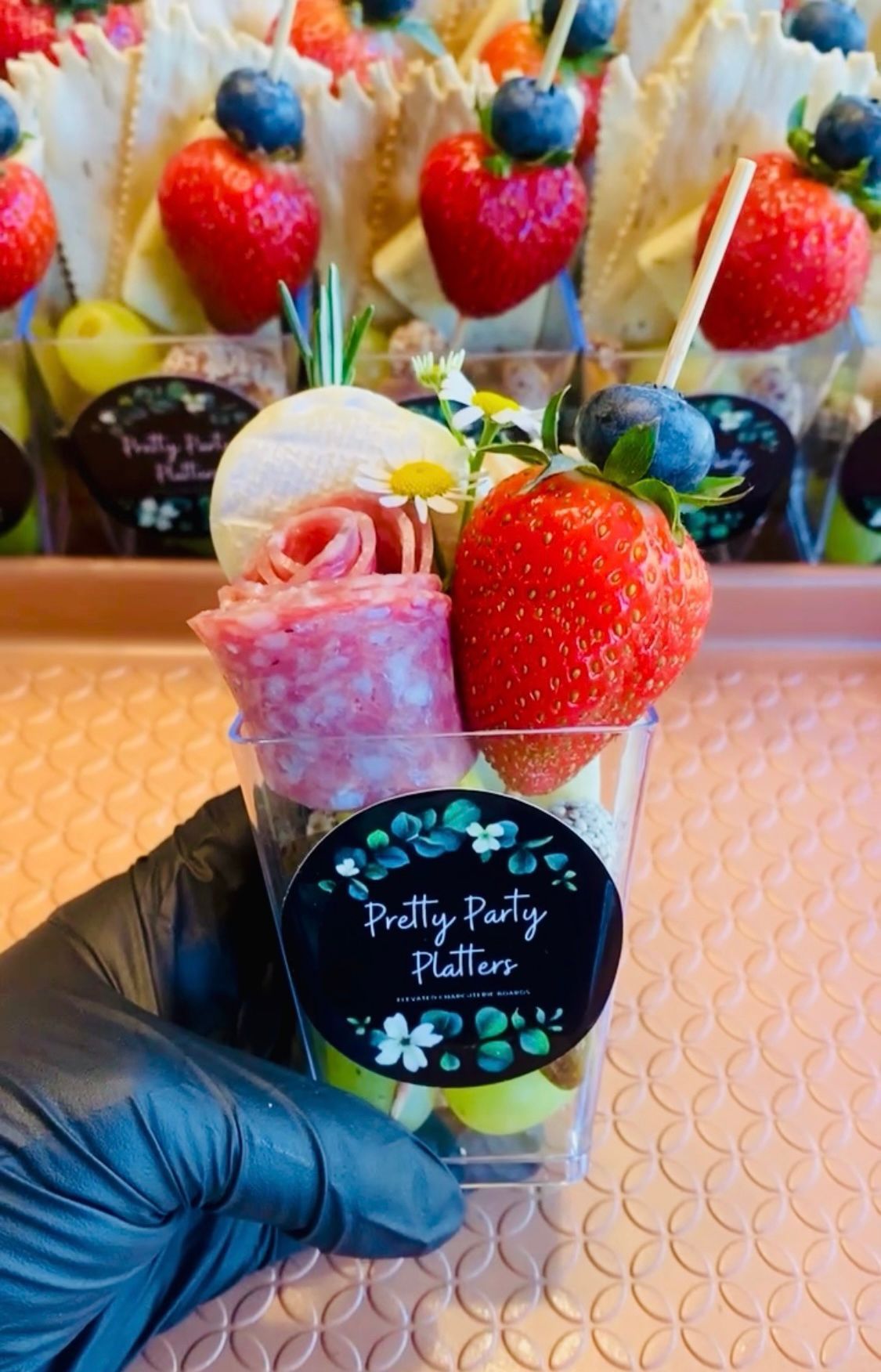 Hand holding a clear cup with fruit, salami, cheese, and a "Pretty Party Platters" sticker.