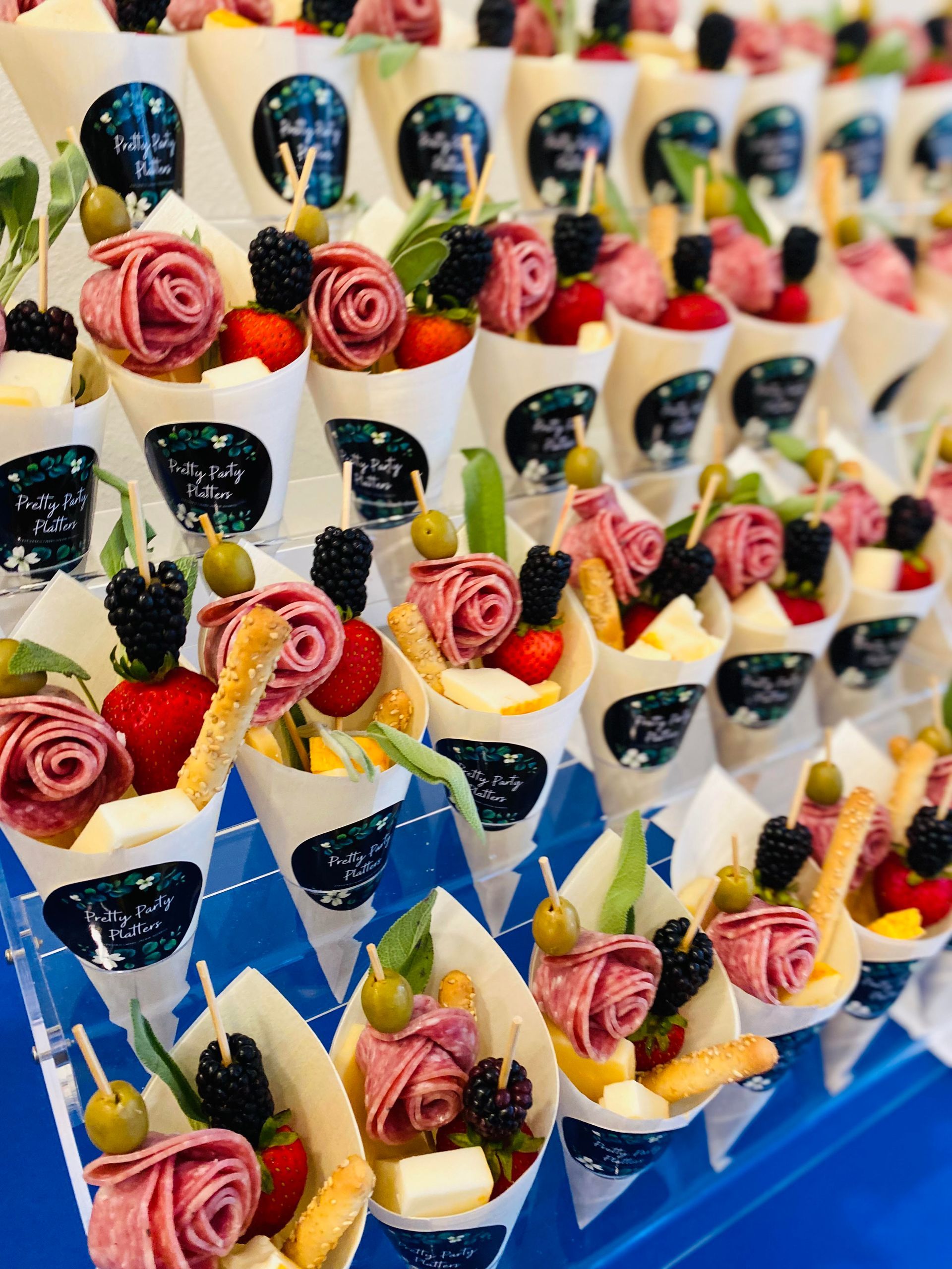Appetizer cones: salami roses, berries, cheese, and olives, arranged in rows on a blue surface.