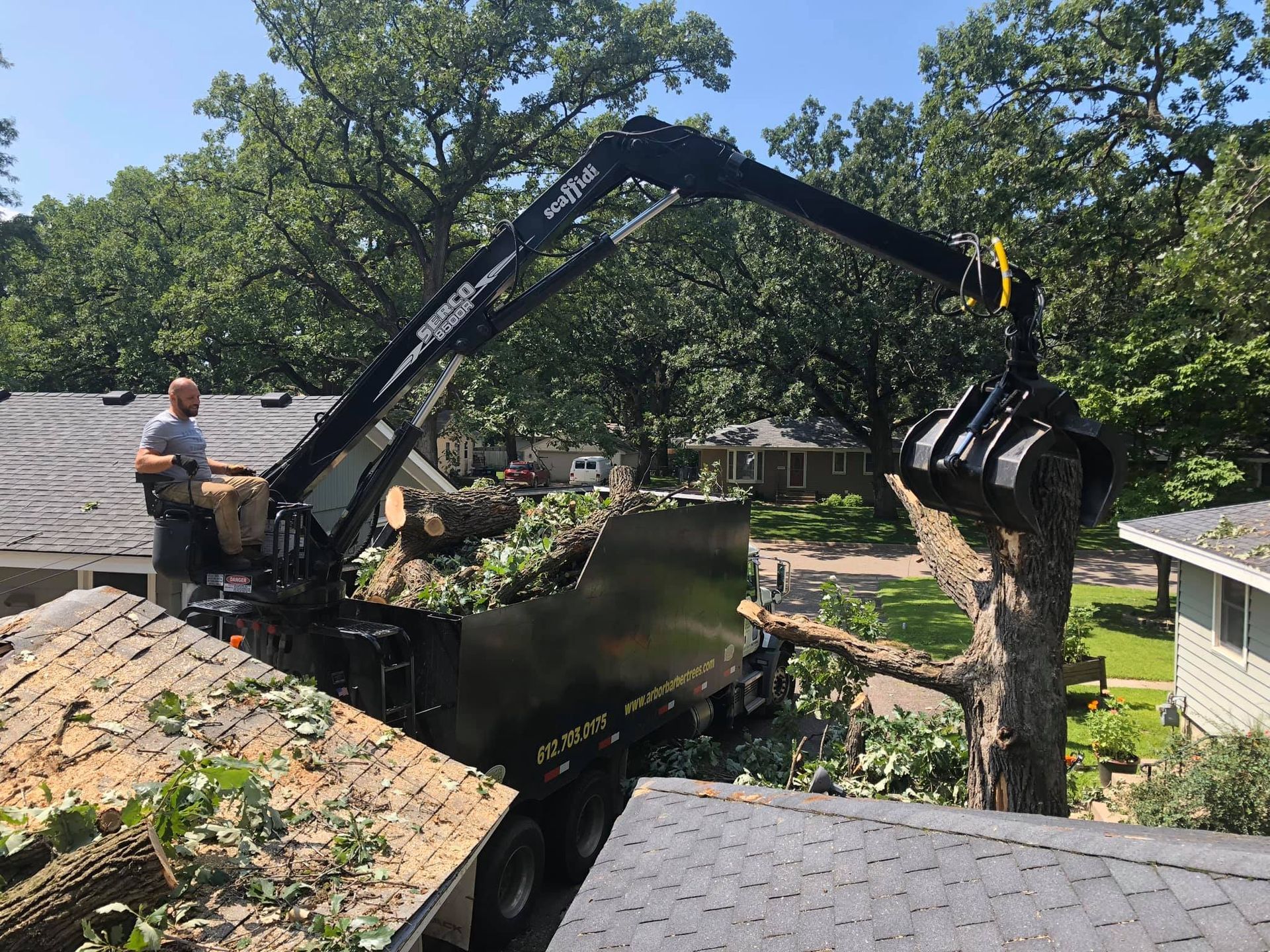 Man operating a crane to load tree branches into a truck from a rooftop on a sunny day.
