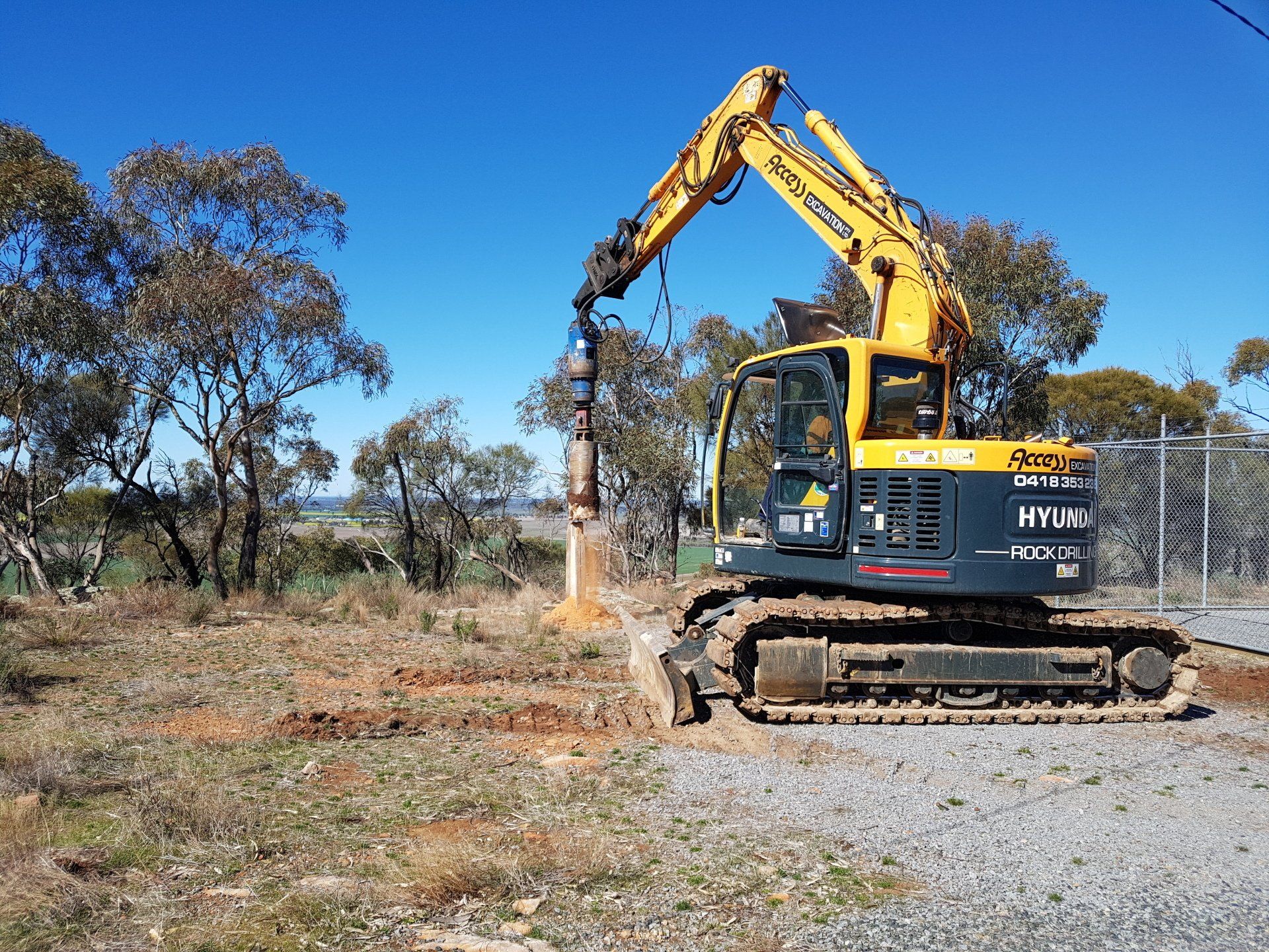 Yellow excavator drilling in a dry field beside trees and a fence
