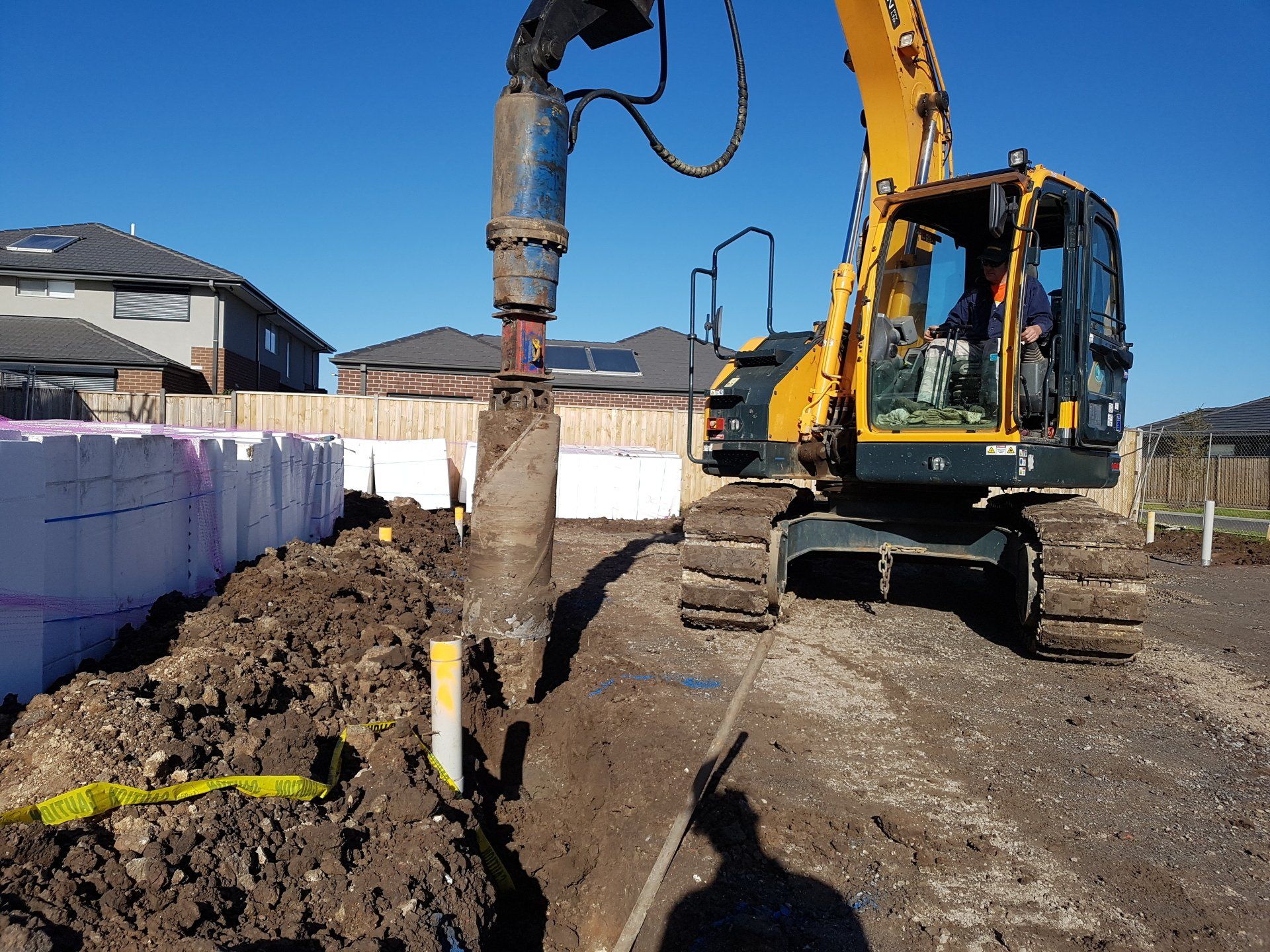 Excavator digging a trench beside a construction site with soil piles and houses in the background