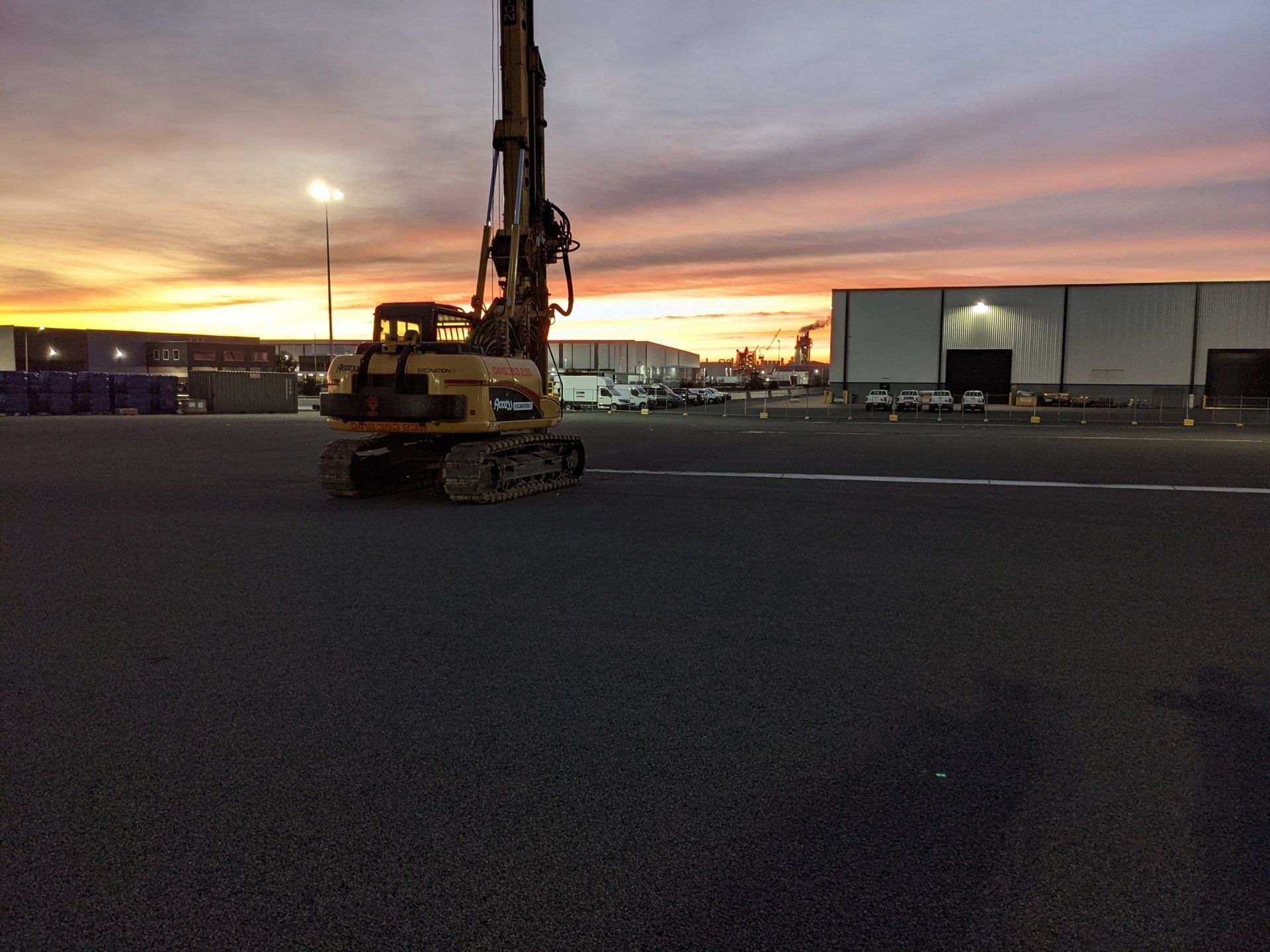 Excavator on a dark lot at sunset, with warehouse buildings and orange sky in the background