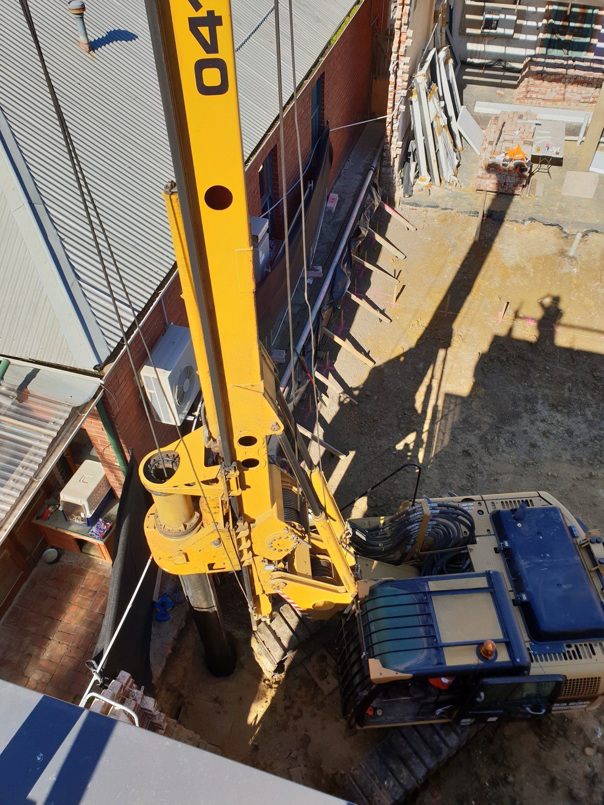 Yellow drilling rig beside a blue truck in a construction site pit, viewed from above