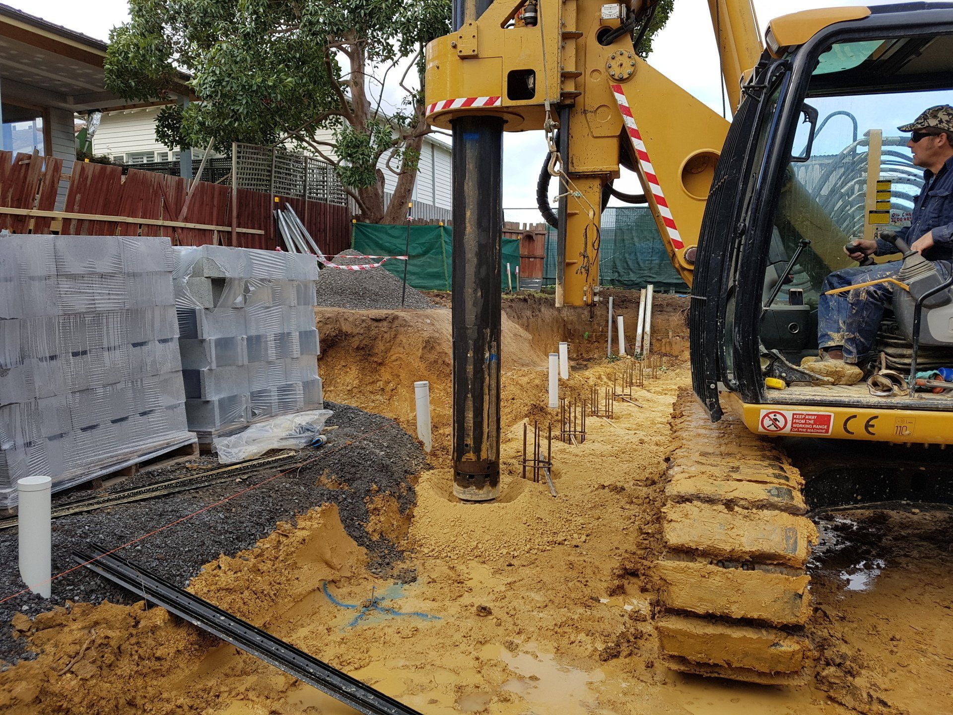 Excavator driving steel piles at a muddy construction site beside stacked concrete blocks
