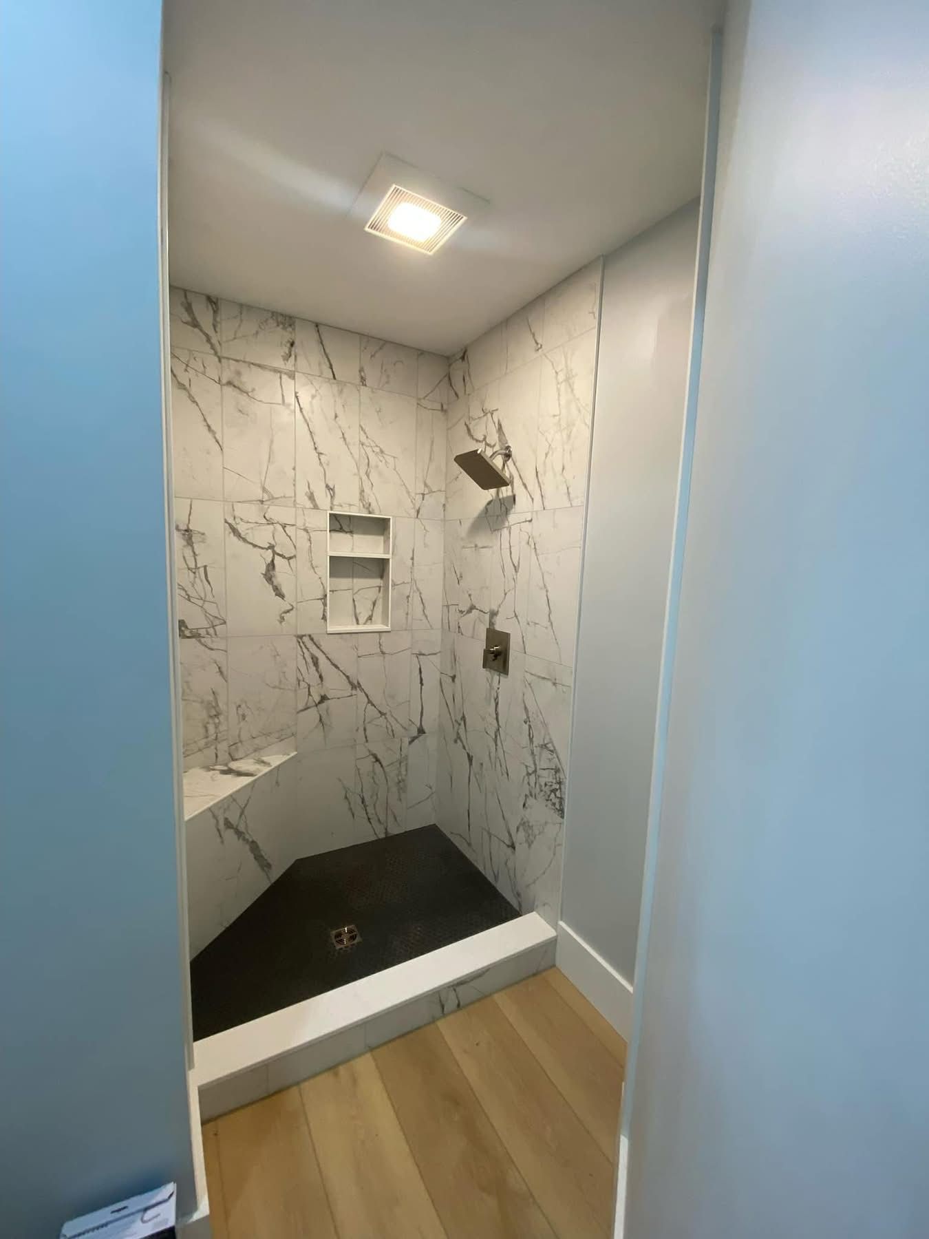 Modern shower with white marble-veined walls, a black tiled floor, a built-in bench, a niche, and a single ceiling light.