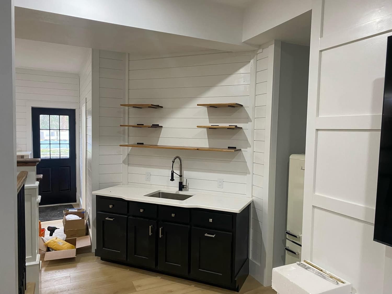 A wet bar with black cabinets, a white countertop, and three wooden floating shelves against a white shiplap wall.