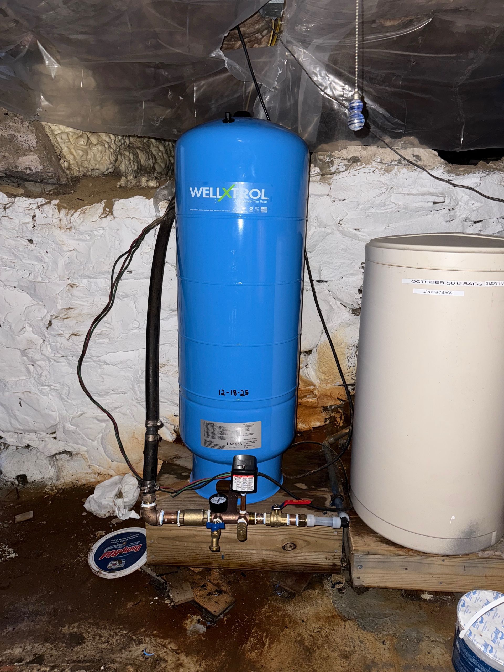A blue Well-Trol well water pressure tank sits on a wooden platform next to a beige tank in a basement crawl space.