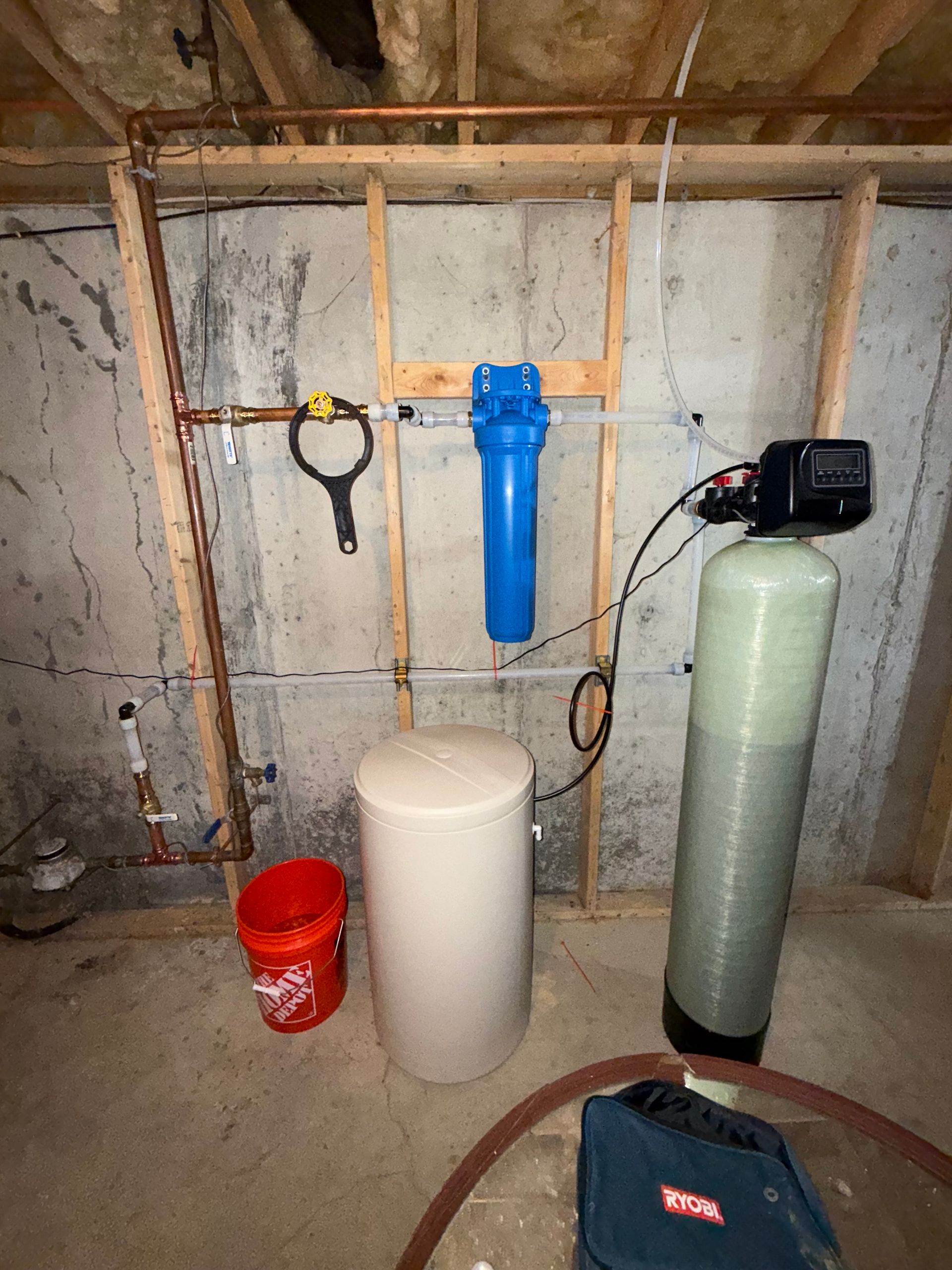 A water filtration and softener system mounted on a basement concrete wall with a blue filter housing and a brine tank.