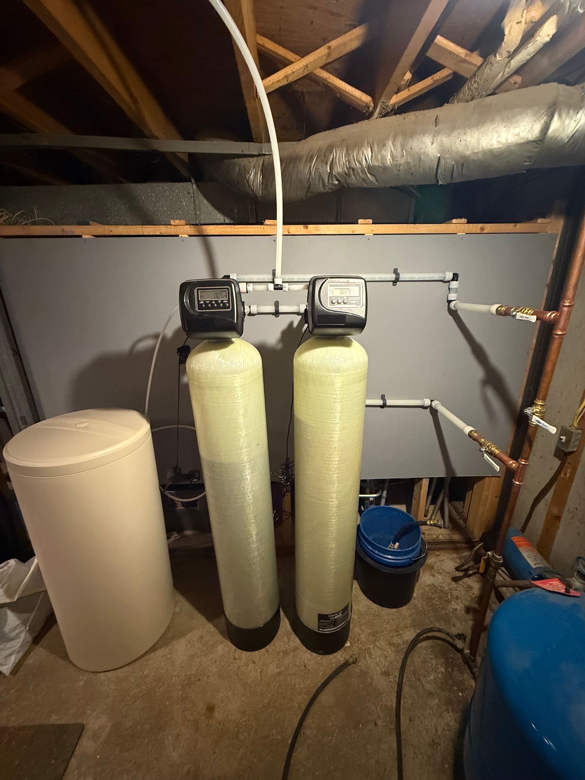 A water treatment system with two tall, beige mineral tanks and control heads, connected by white pipes in a basement.