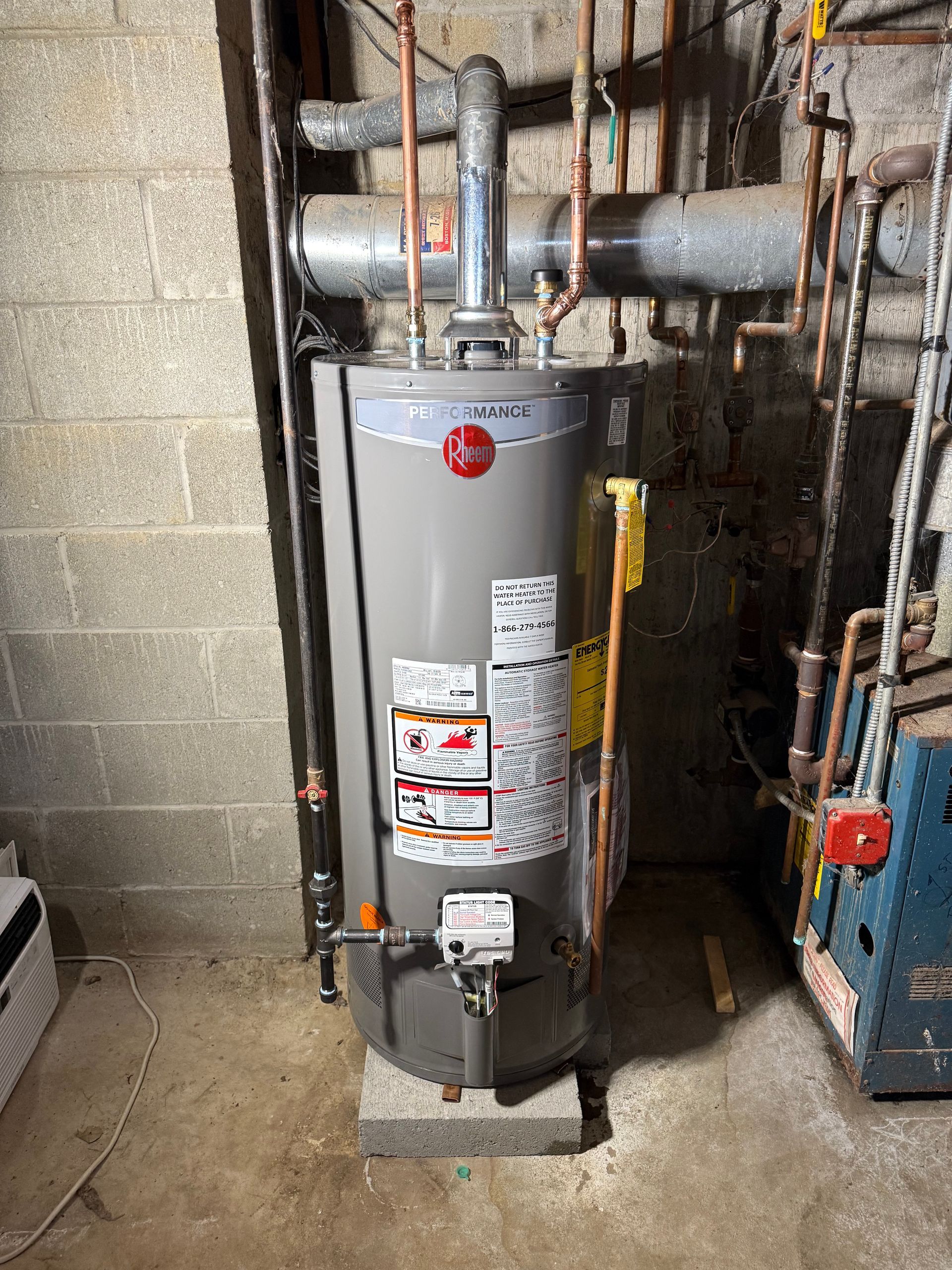 A grey Rheem gas water heater stands on a concrete block in a basement next to copper piping and a cinder block wall.