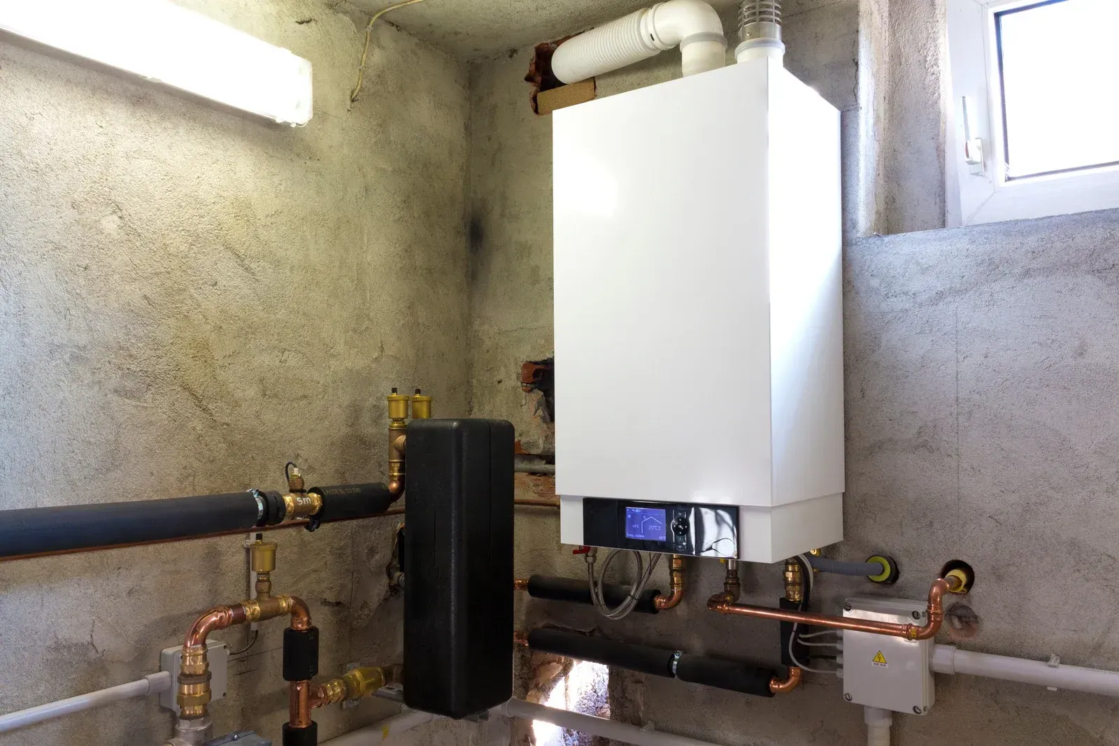 A close-up view of a white water heater tank with various metal pipes, valves, and electrical grounding wires attached.