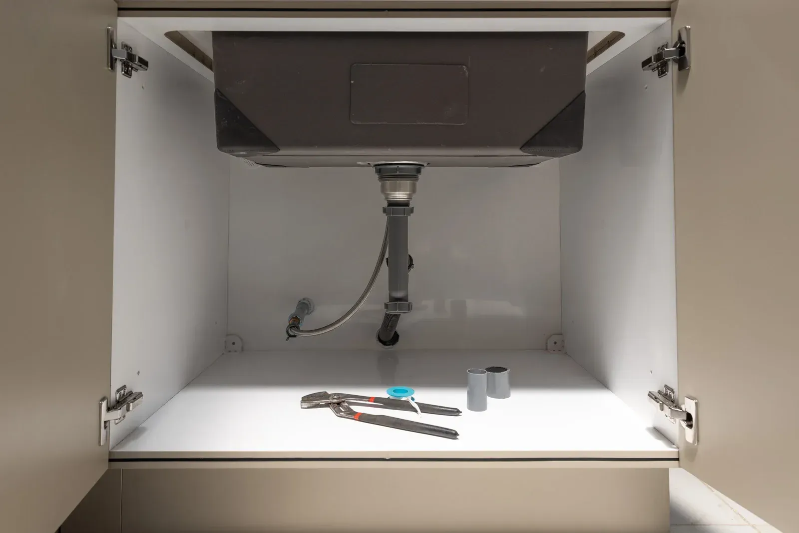 Pliers and plumbing components sit on the base of an open kitchen cabinet beneath a sink drain assembly.