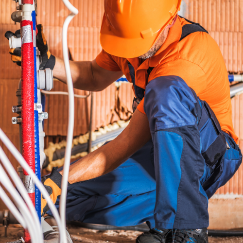 A worker in an orange hard hat and shirt with blue work pants connects plumbing pipes in a building under construction.