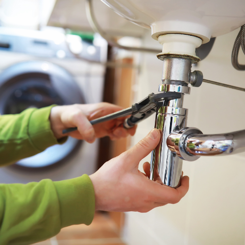 A person wearing a green sweater uses a wrench to tighten a chrome P-trap pipe under a sink.