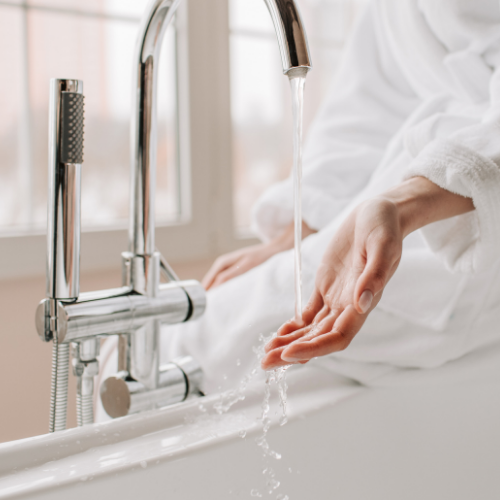A person wearing a white bathrobe reaches their hand under a running bathroom faucet.