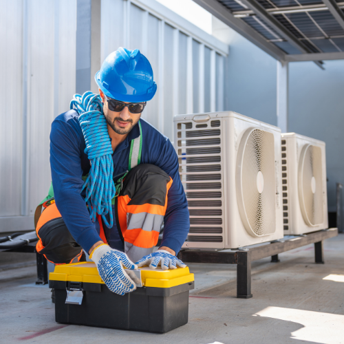 A technician in a blue hard hat, sunglasses, and safety gear kneeling to open a toolbox near two outdoor AC units.