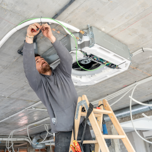 A technician on a ladder wiring a ceiling-mounted HVAC unit inside an unfinished building.