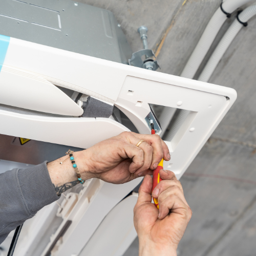 A person uses a screwdriver to tighten a metal bracket on a white, ceiling-mounted indoor HVAC unit.