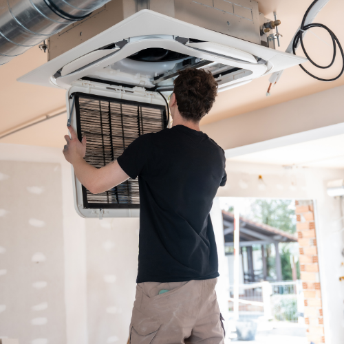 A person in a black shirt and tan pants installs a square air filter into a white ceiling-mounted HVAC unit.