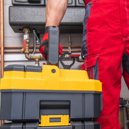 A worker in red coveralls and work gloves carries a yellow and black tool box in front of mechanical pipes.