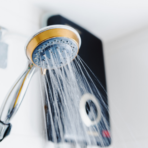 A hand-held shower head with a gold-colored rim sprays water, with a black electric water heater unit in the background.