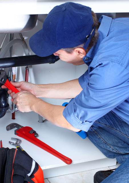 A plumber in a blue uniform and cap using a red pipe wrench to fix pipes under a kitchen sink.