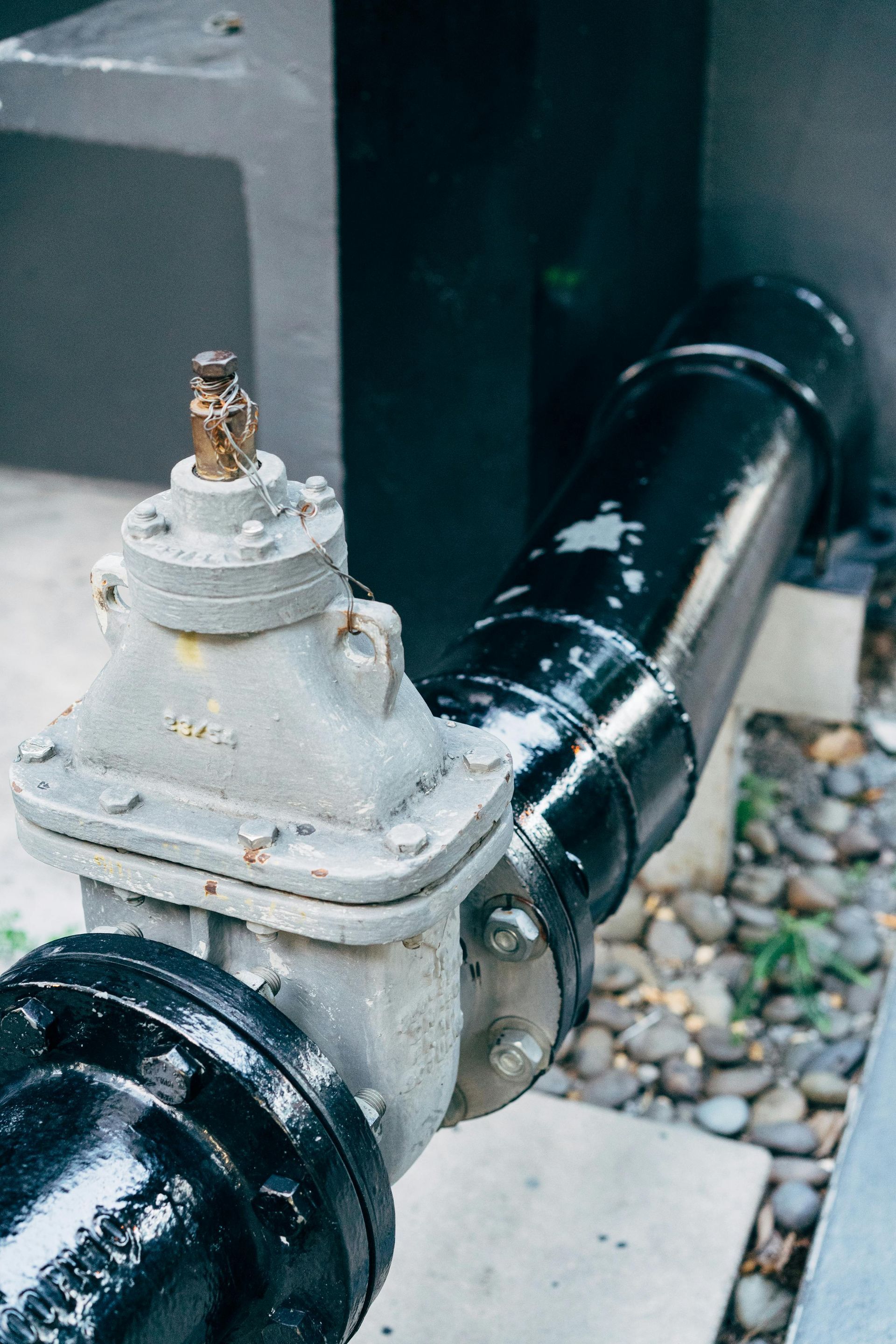 A gray industrial valve connected to a black metal pipe outdoors, set against a concrete base and gravel.