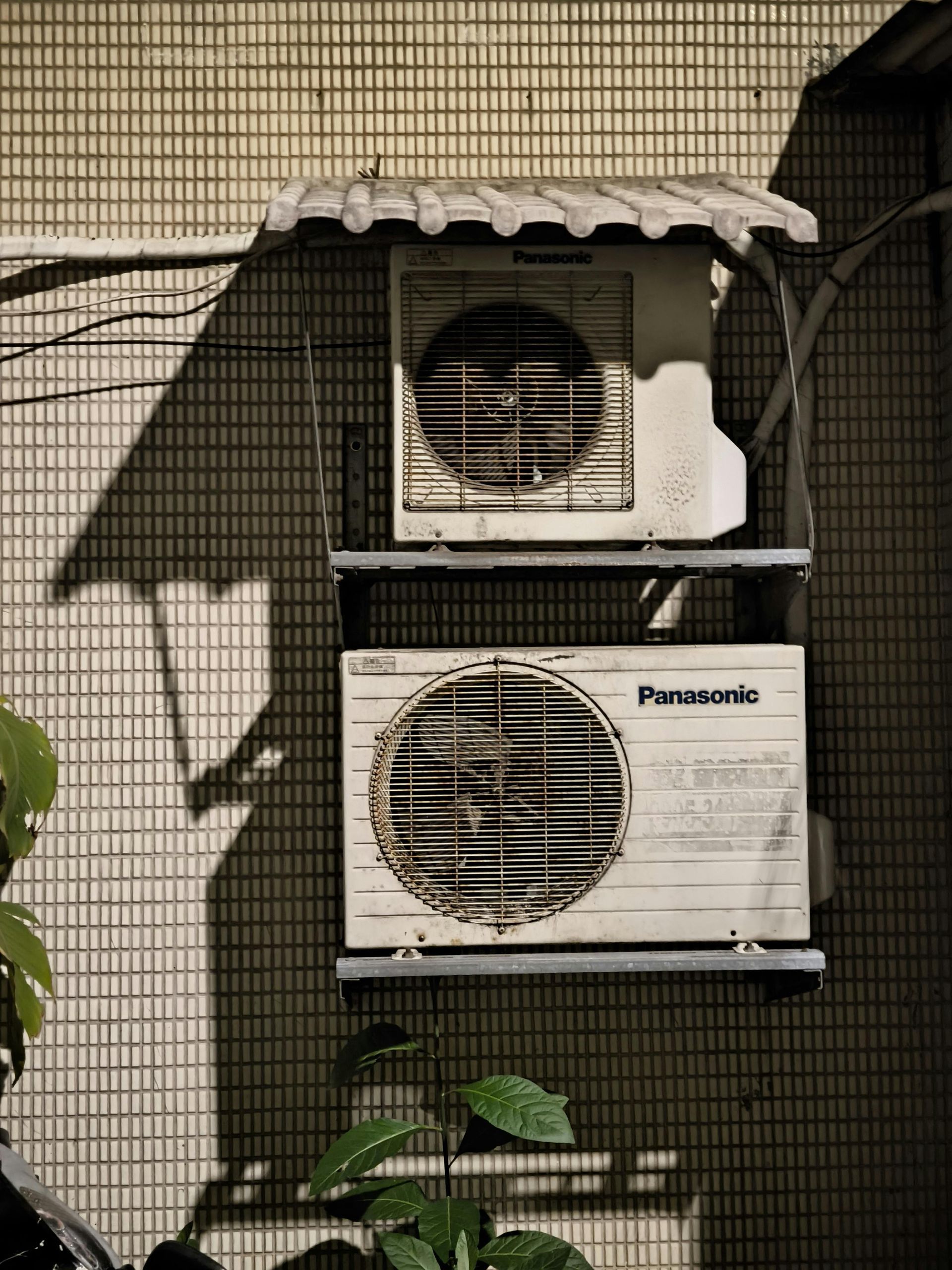 Two white Panasonic air conditioner units stacked vertically on a metal bracket against a tiled wall.