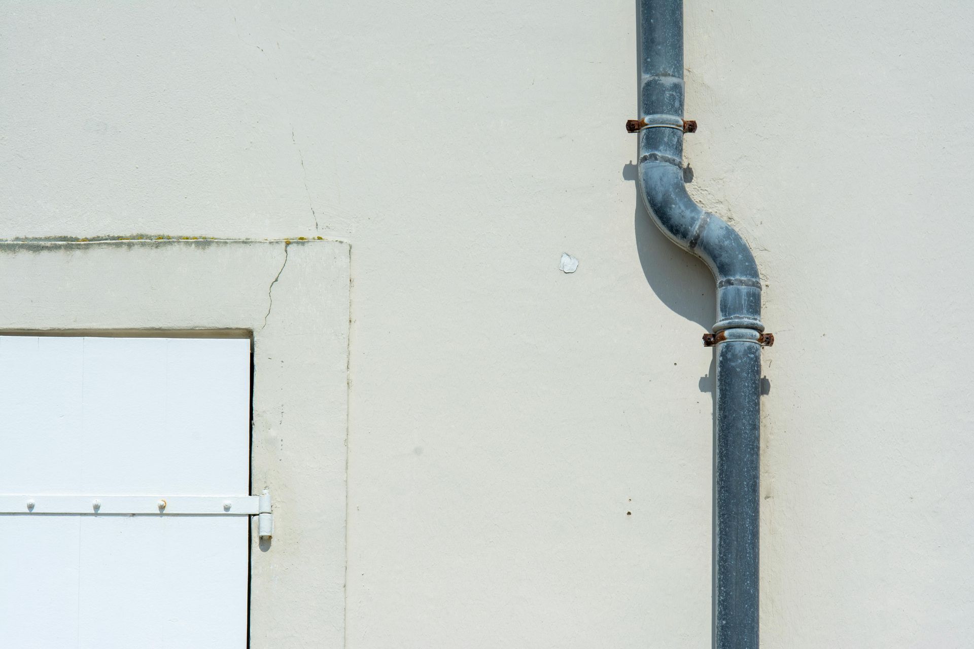 A vertical drainpipe with a small offset curve runs down a smooth, off-white exterior wall next to a window frame.