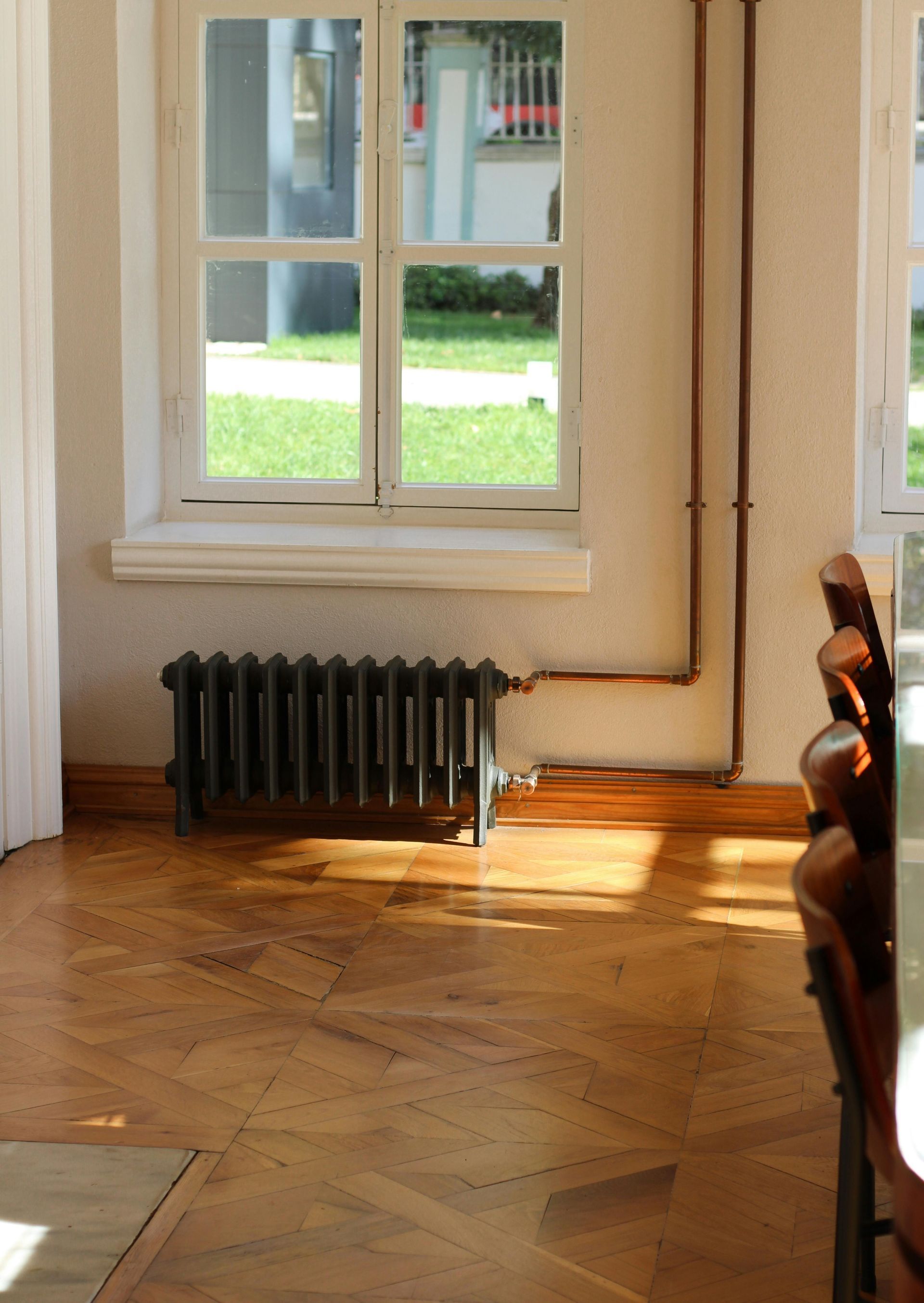 A dark-colored cast iron radiator sits beneath a window in a room with hardwood parquet flooring and visible pipework.
