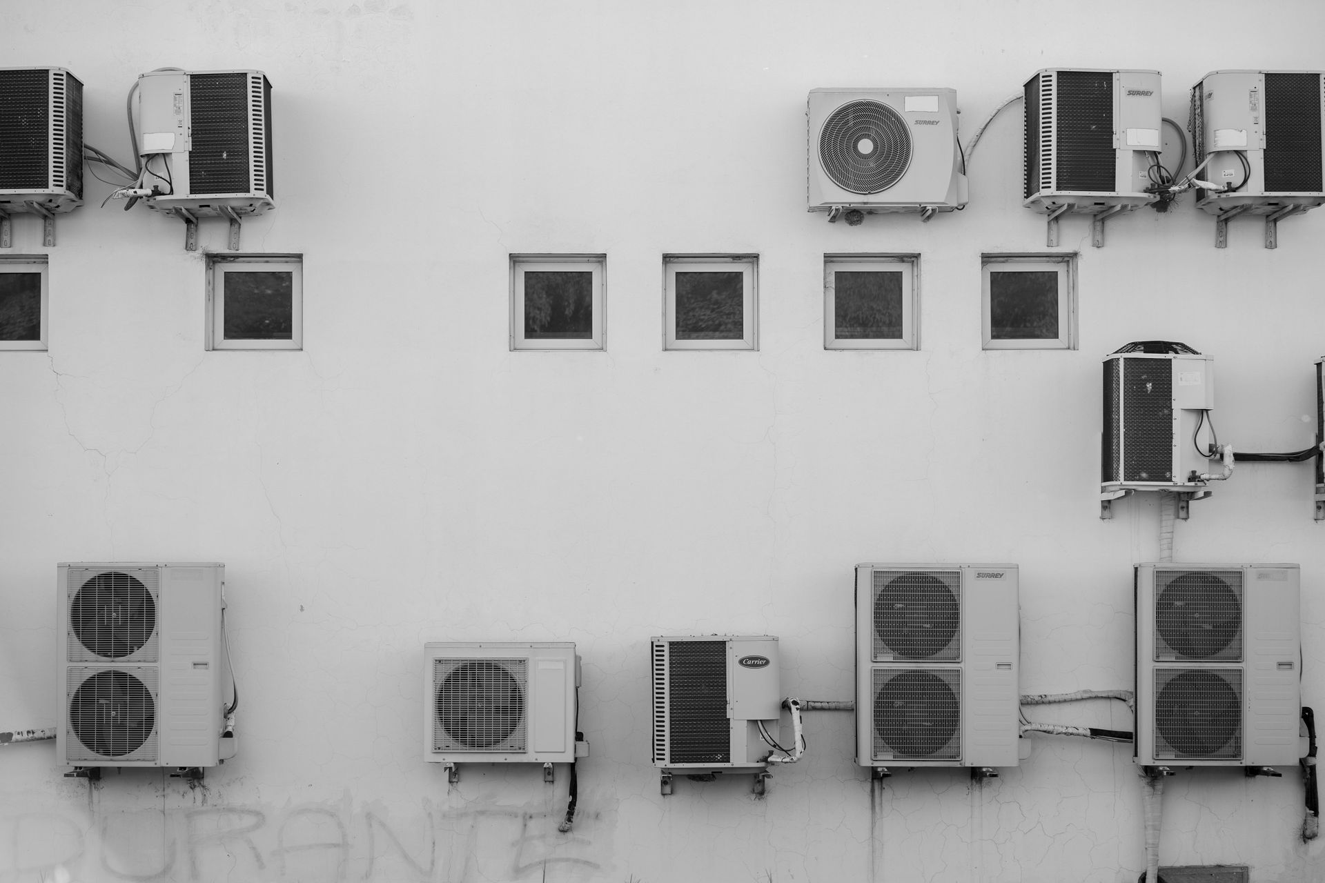 Multiple air conditioning units mounted on a plain exterior wall with several small, square windows.
