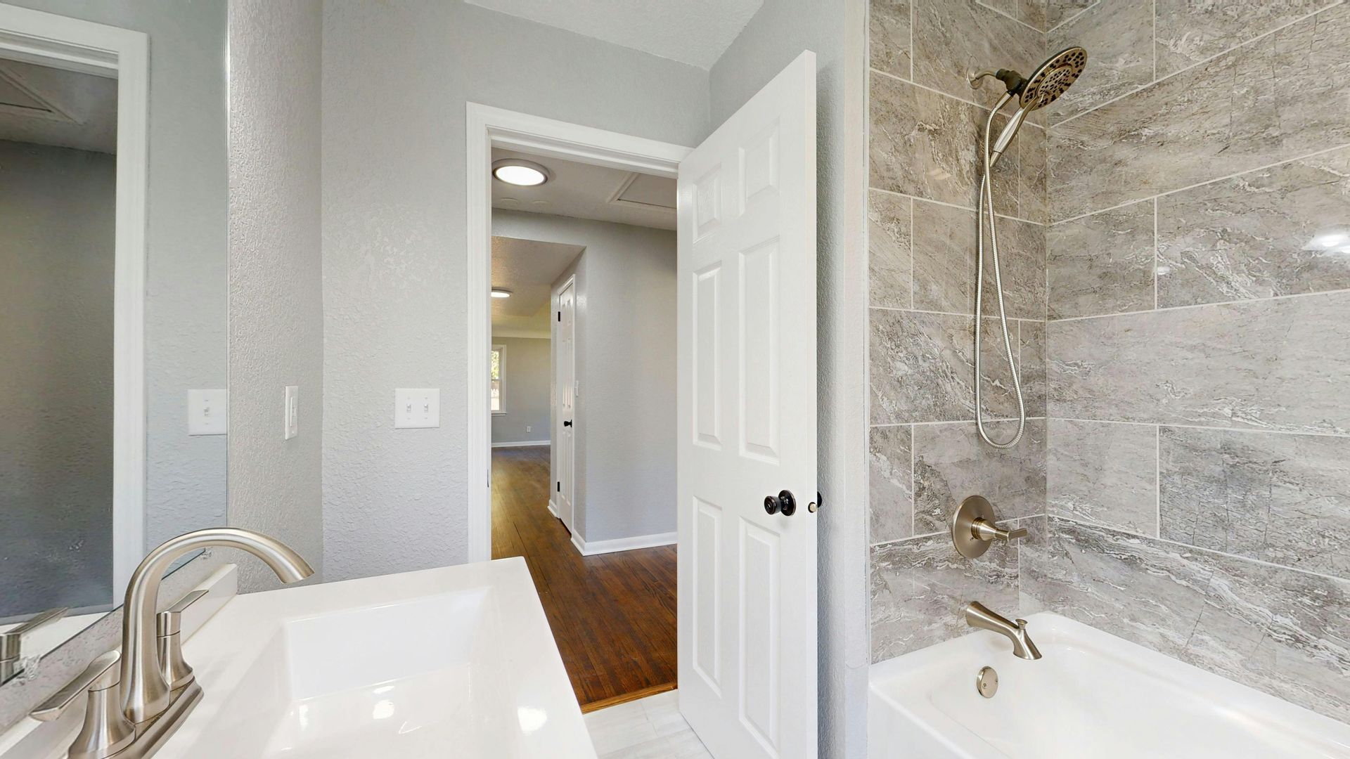 A bathroom with a white vanity, a tiled tub and shower, and an open door leading to a hallway with wood-style flooring.