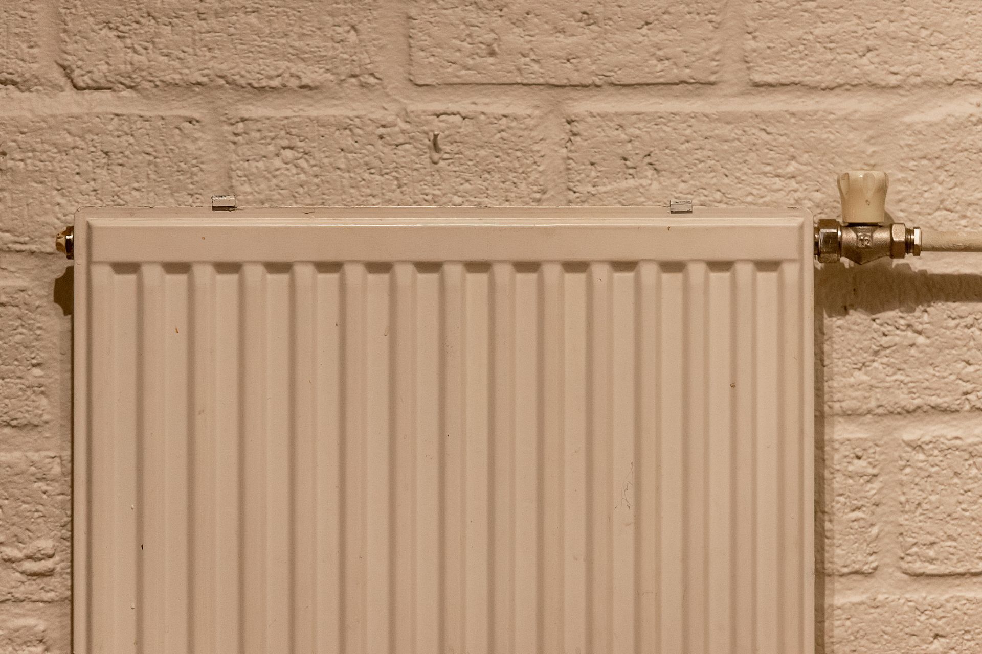 A white, ribbed radiator mounted on a textured, off-white brick wall, featuring a manual valve on the right side.