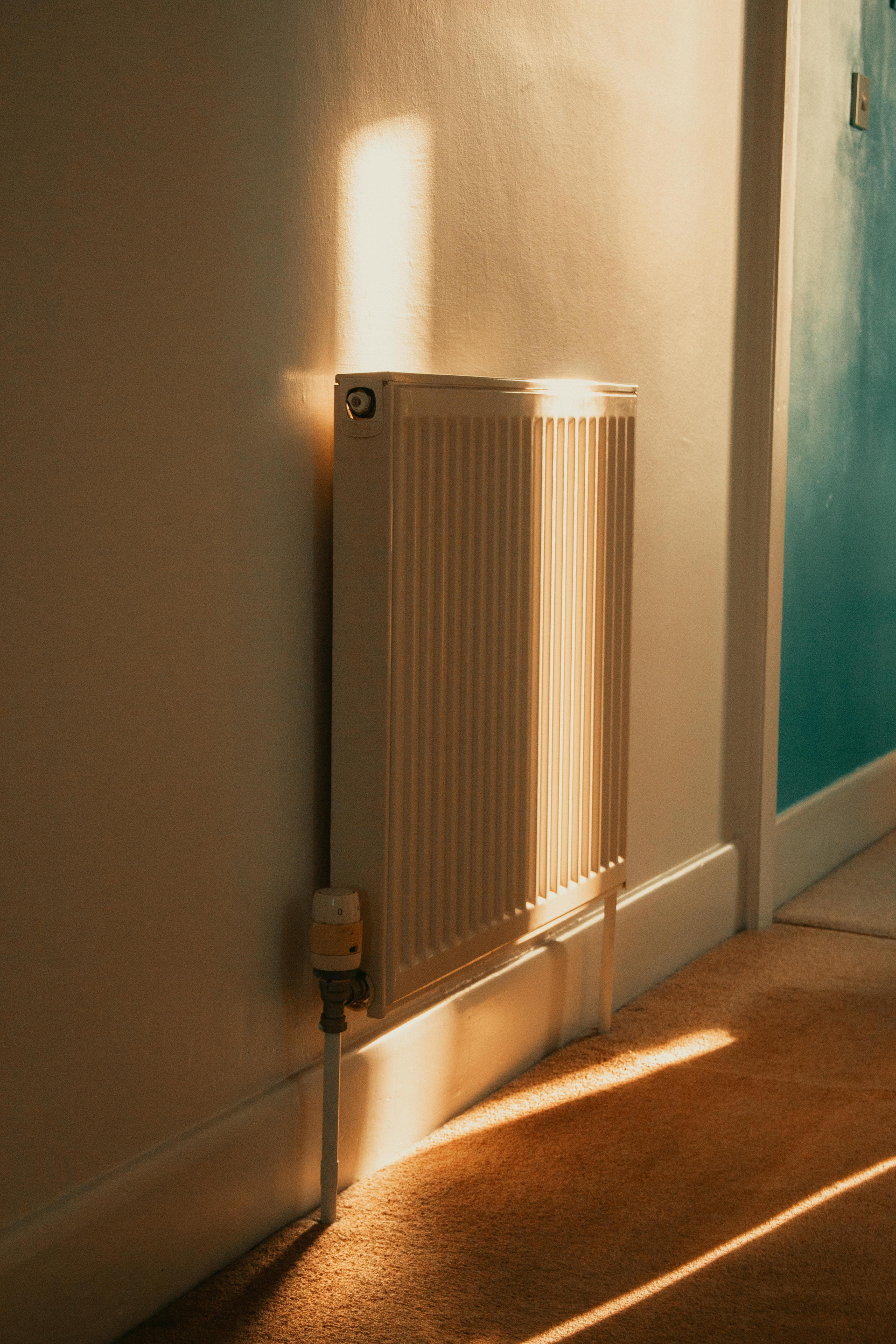 A vertical white radiator mounted on a wall in a room illuminated by warm, slanting sunlight.