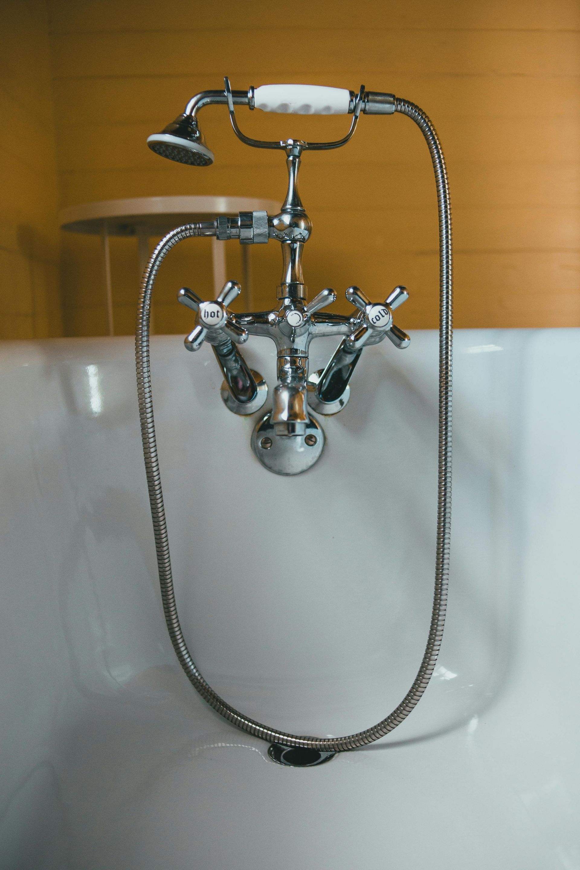 A chrome bathtub faucet with a flexible handheld showerhead attached to a white tub against a yellow wall.