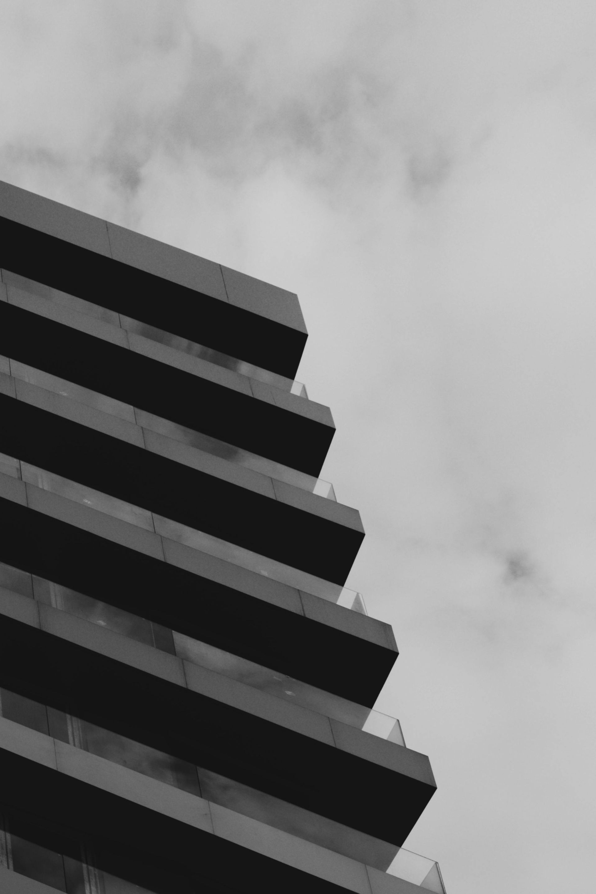 Low-angle, black-and-white view of the corner of a modern high-rise building with repeating balconies against a cloudy sky.