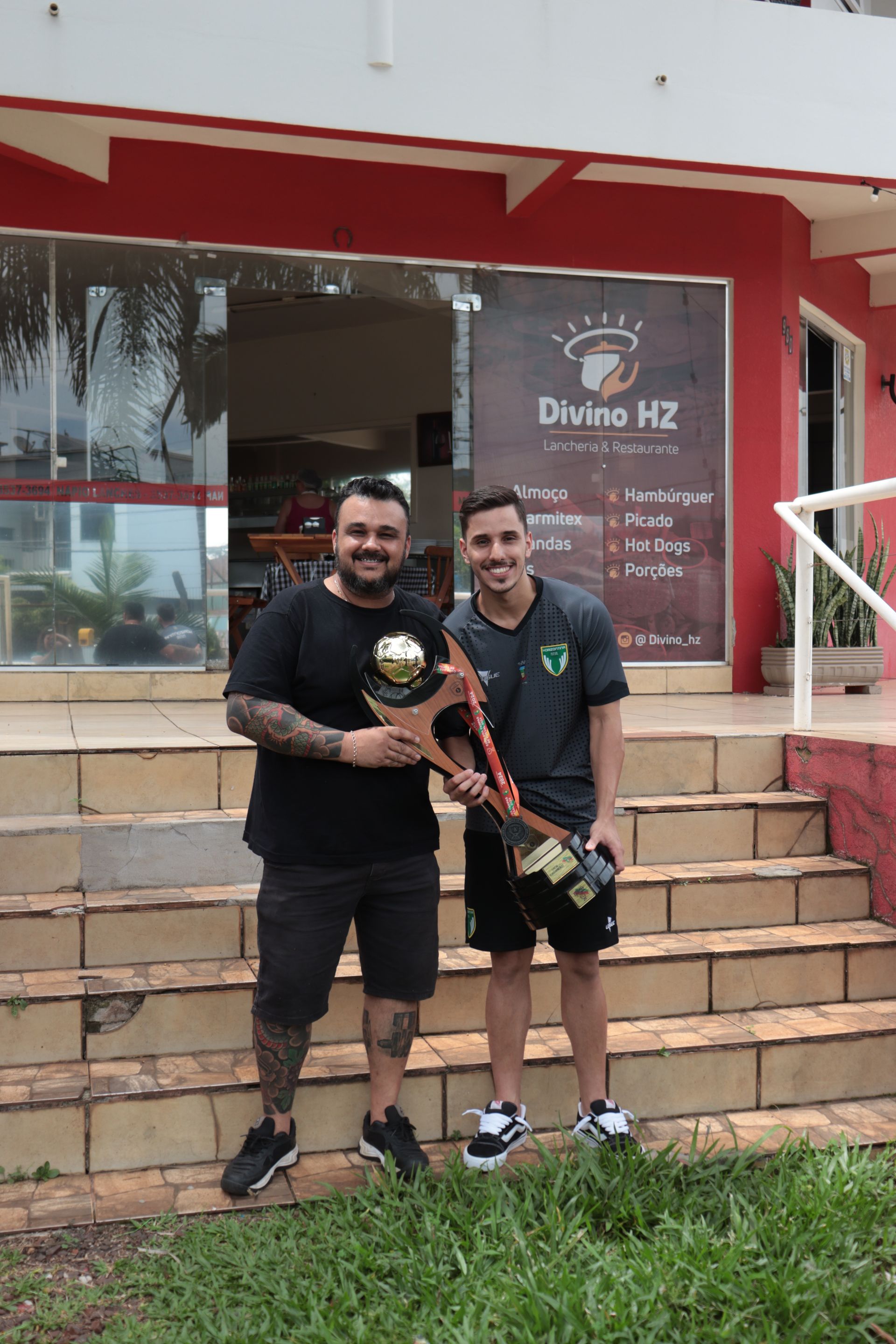 Dois homens estão parados em frente a um prédio segurando troféus.