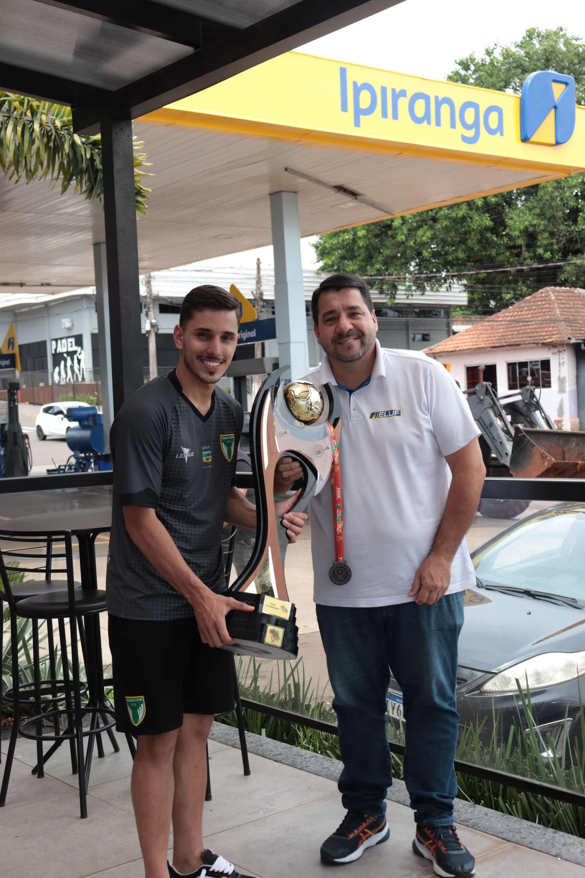 Dois homens estão de pé, um ao lado do outro, segurando um troféu.