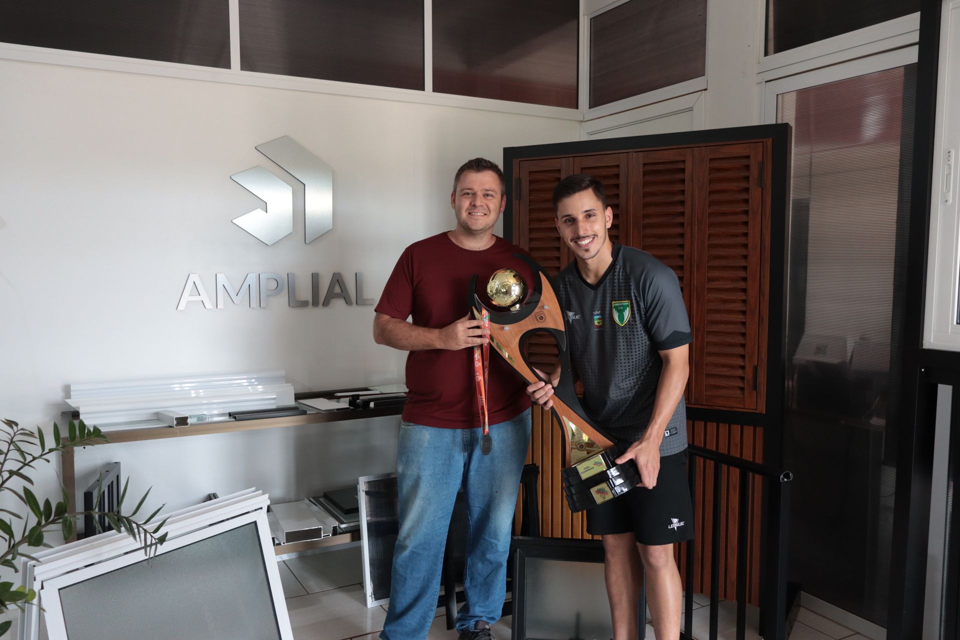 Dois homens estão de pé, um ao lado do outro, em uma sala, segurando um troféu.