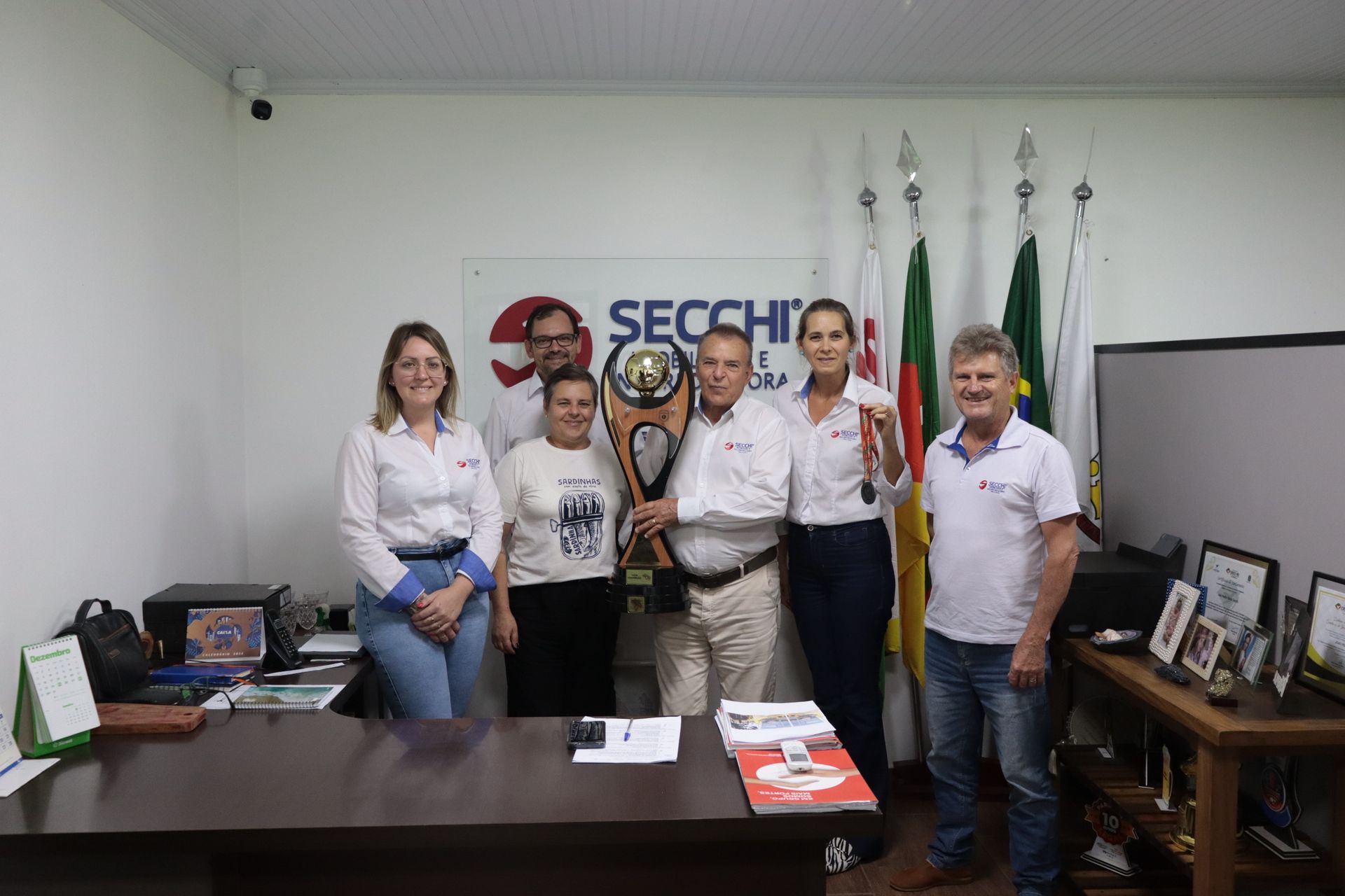 Um grupo de pessoas está em pé ao redor de uma mesa em uma sala segurando um troféu.