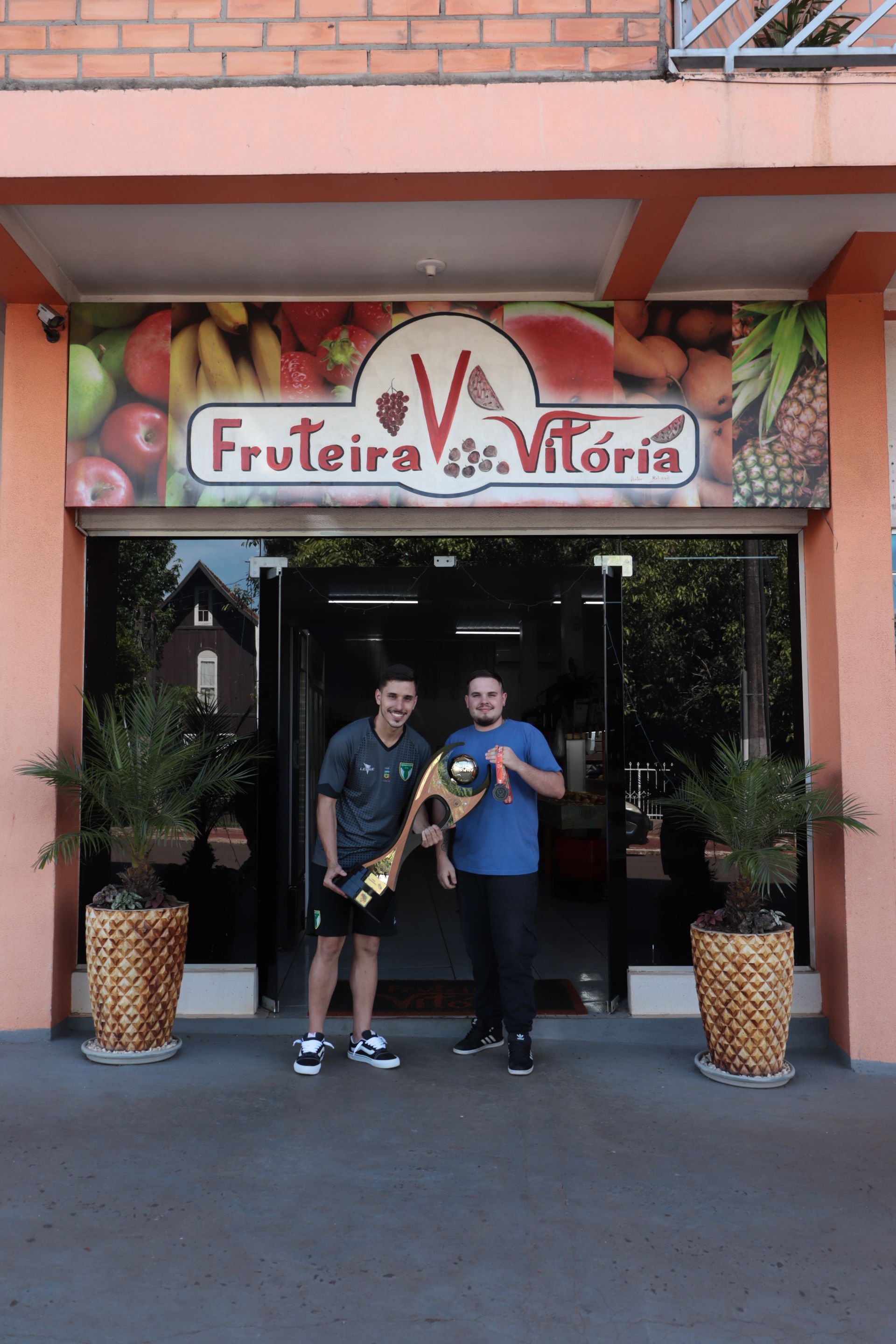 Dois homens parados em frente a uma loja chamada Fruteira Vitória