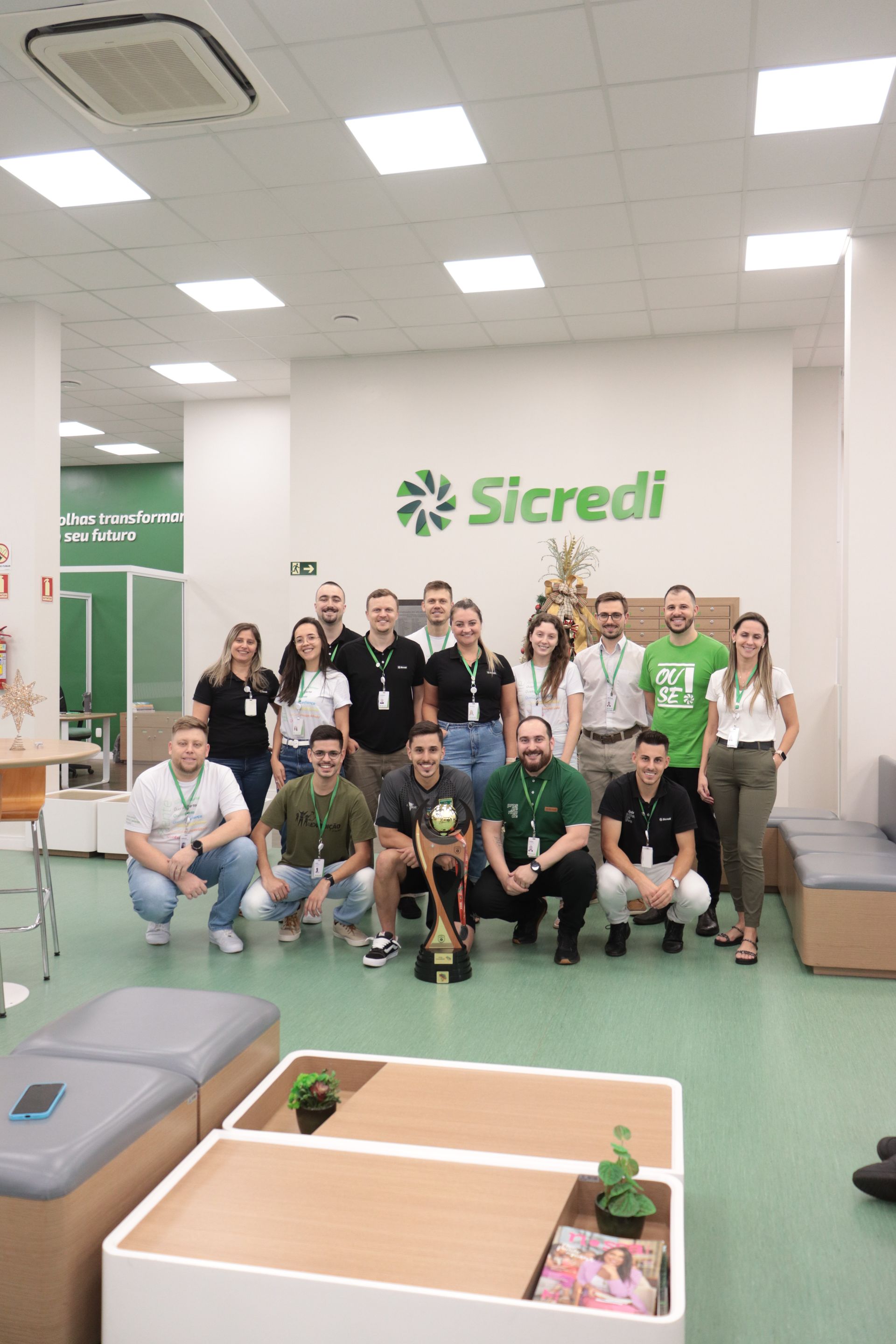 Um grupo de pessoas está posando para uma foto em frente a uma placa do Sicredi.