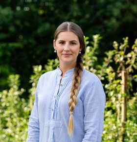 Eine Frau in einem blauen Hemd mit Zopf steht vor einem Busch.