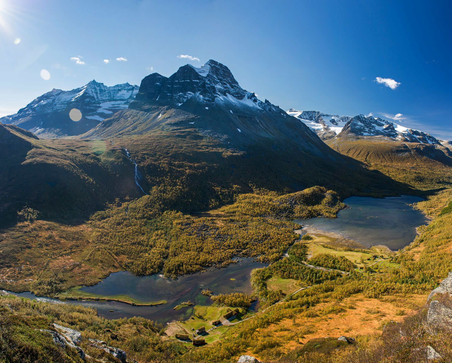 Een panoramisch uitzicht op een met sneeuw bedekte bergtop die boven een vallei uitsteekt met levendige herfstbomen en diepblauwe meren.