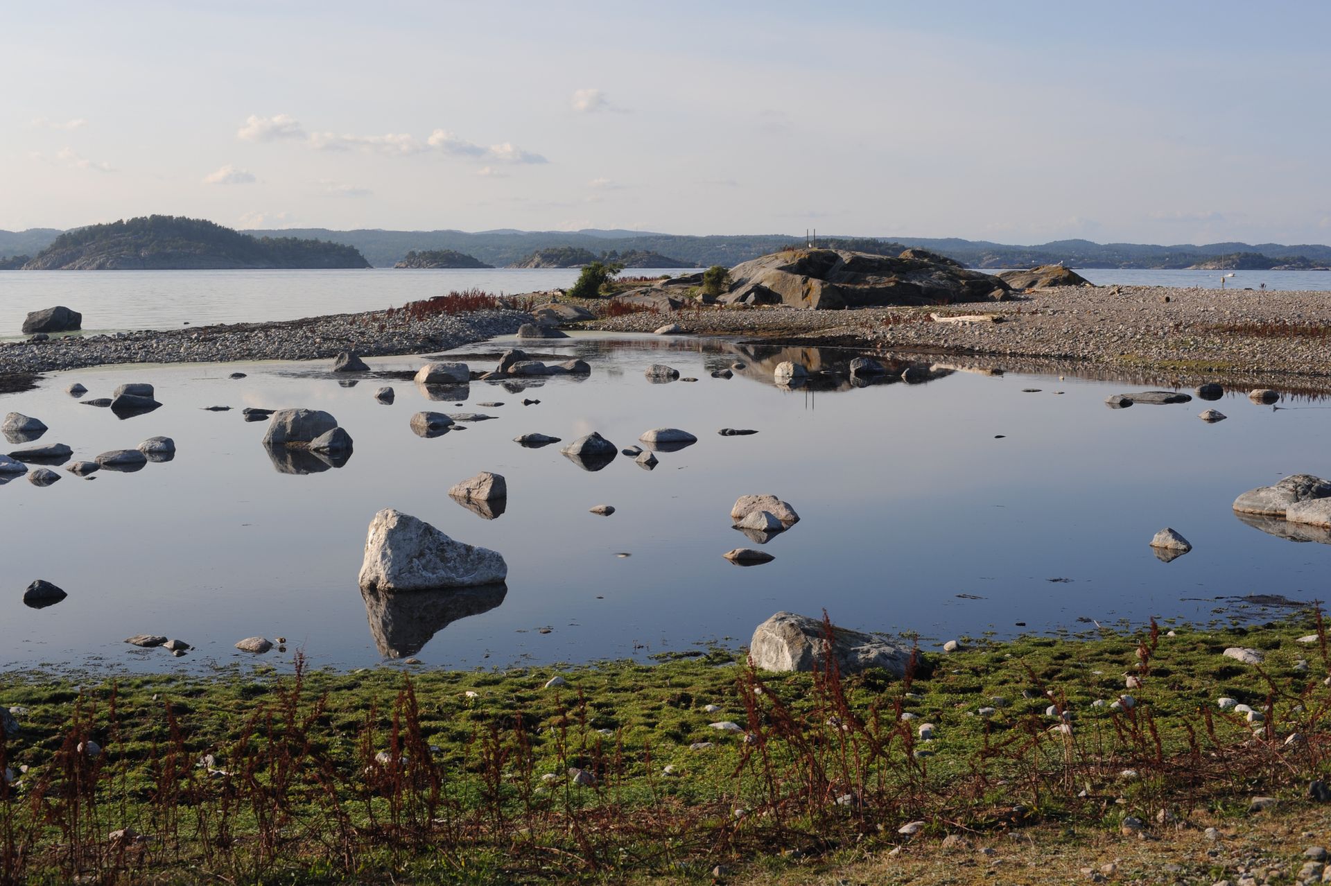 Een rotsachtige kustlijn met een rustig getijdenmeer op de voorgrond, dat overgaat in een baai met eilanden in de verte onder een heldere hemel.