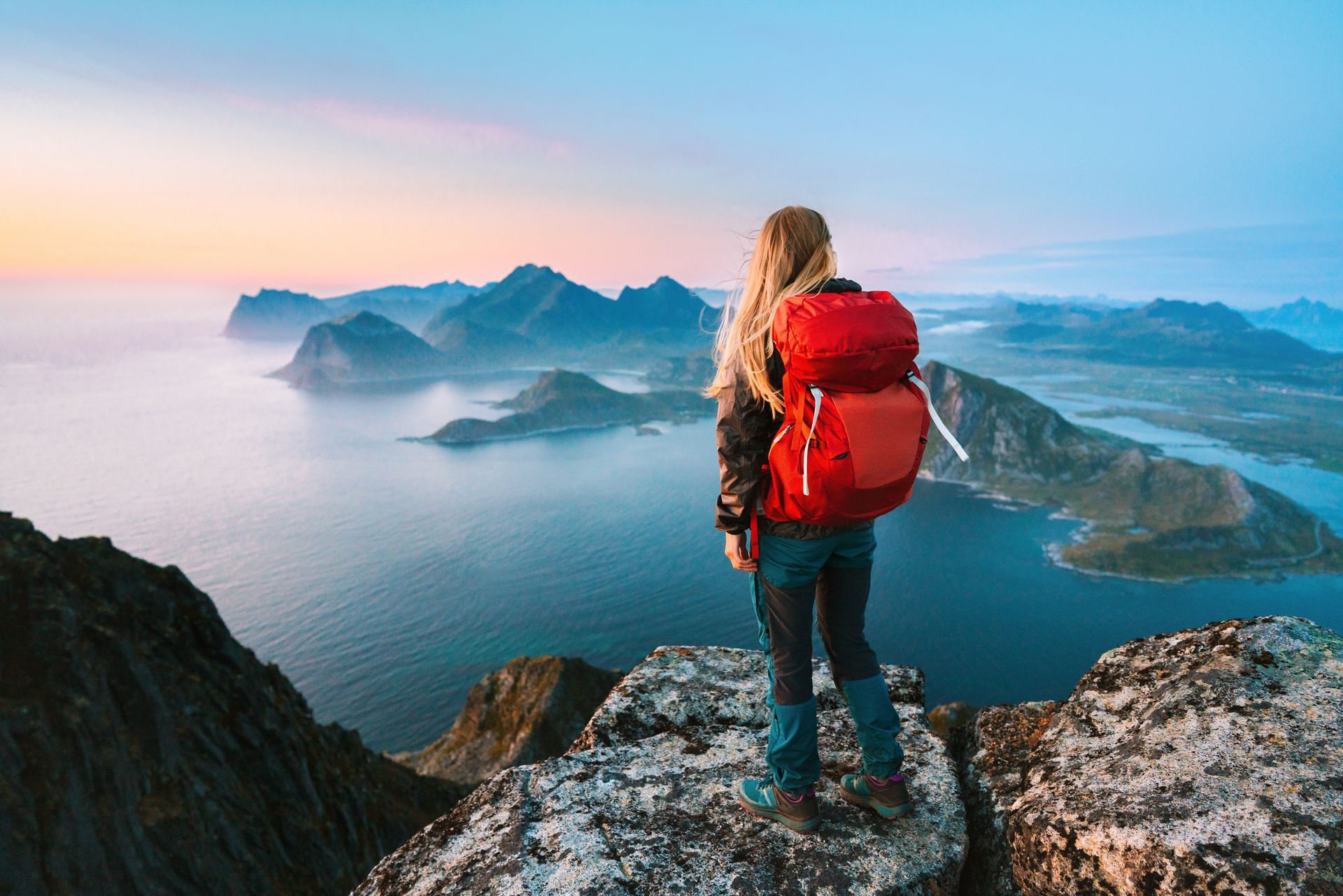 Een wandelaar met een rode rugzak staat op een rotsachtige bergtop en kijkt uit over een schilderachtig landschap met oceaan en eilanden bij zonsondergang.